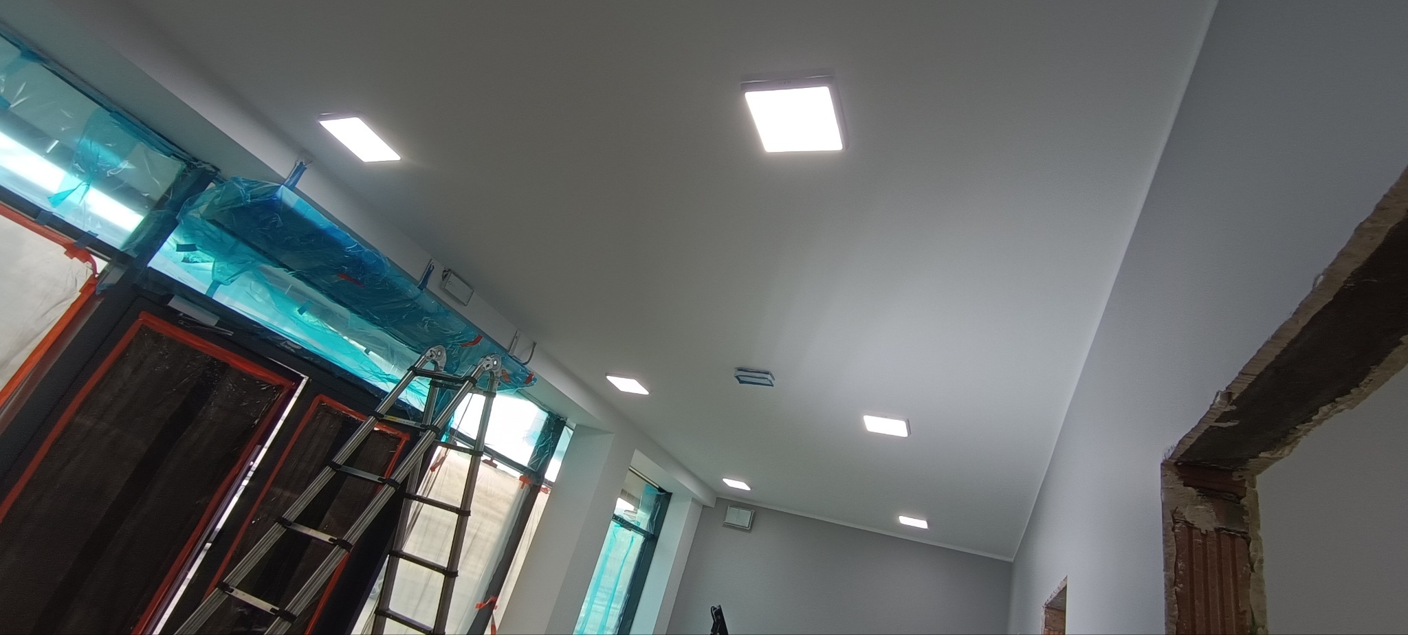 Biały sufit z zamontowanymi, kwadratowymi lampami LED. Widoczne okna zabezpieczone folią i drabina. Ujęcie z dołu, podkreślające nowoczesne oświetlenie.