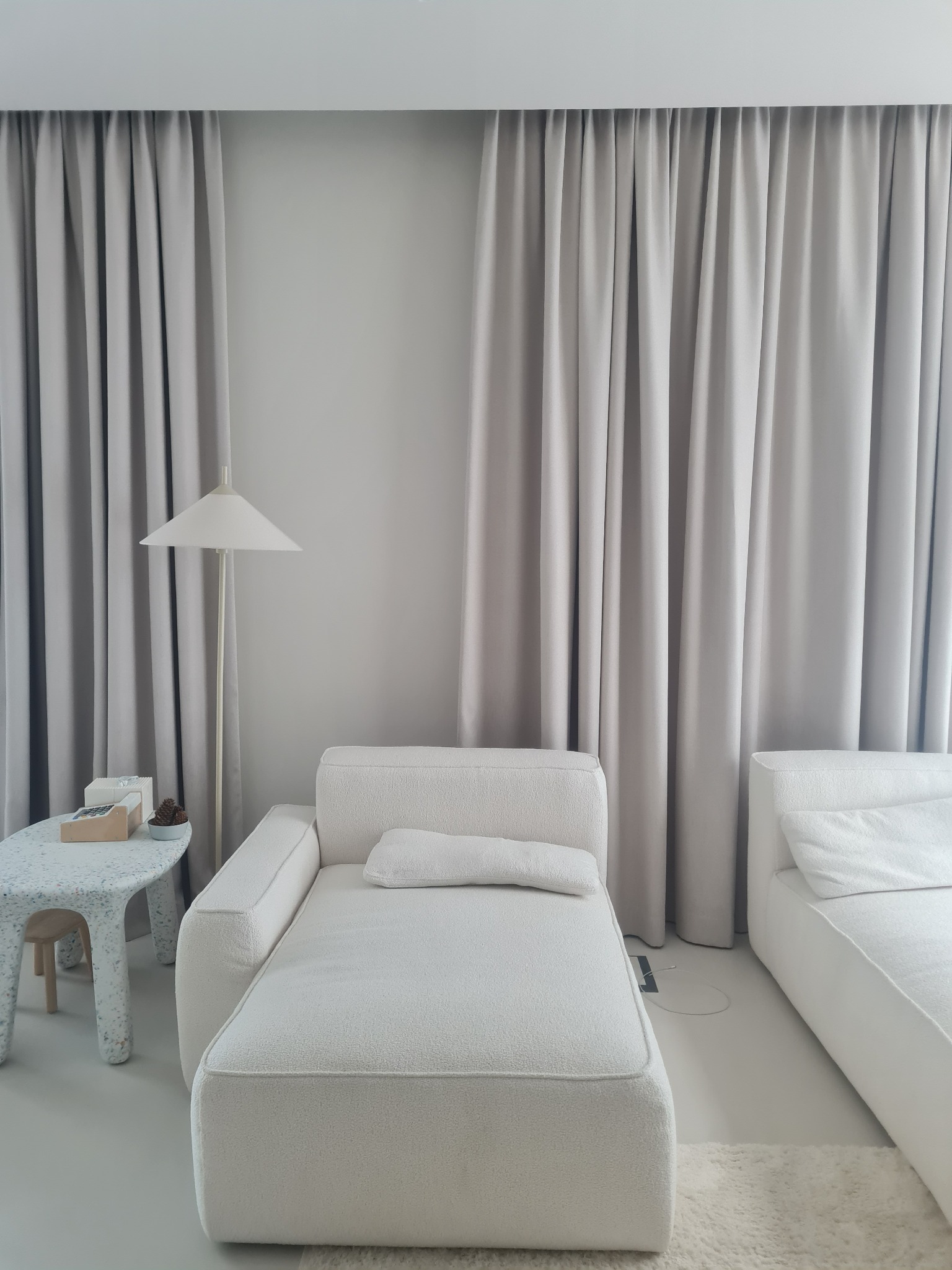 Minimalistyczne wnętrze z beżowymi zasłonami, białymi sofami z miękką tapicerką i stolikiem o nieregularnym kształcie z plamkami, oświetlone lampą podłogową z kloszem w kształcie stożka.