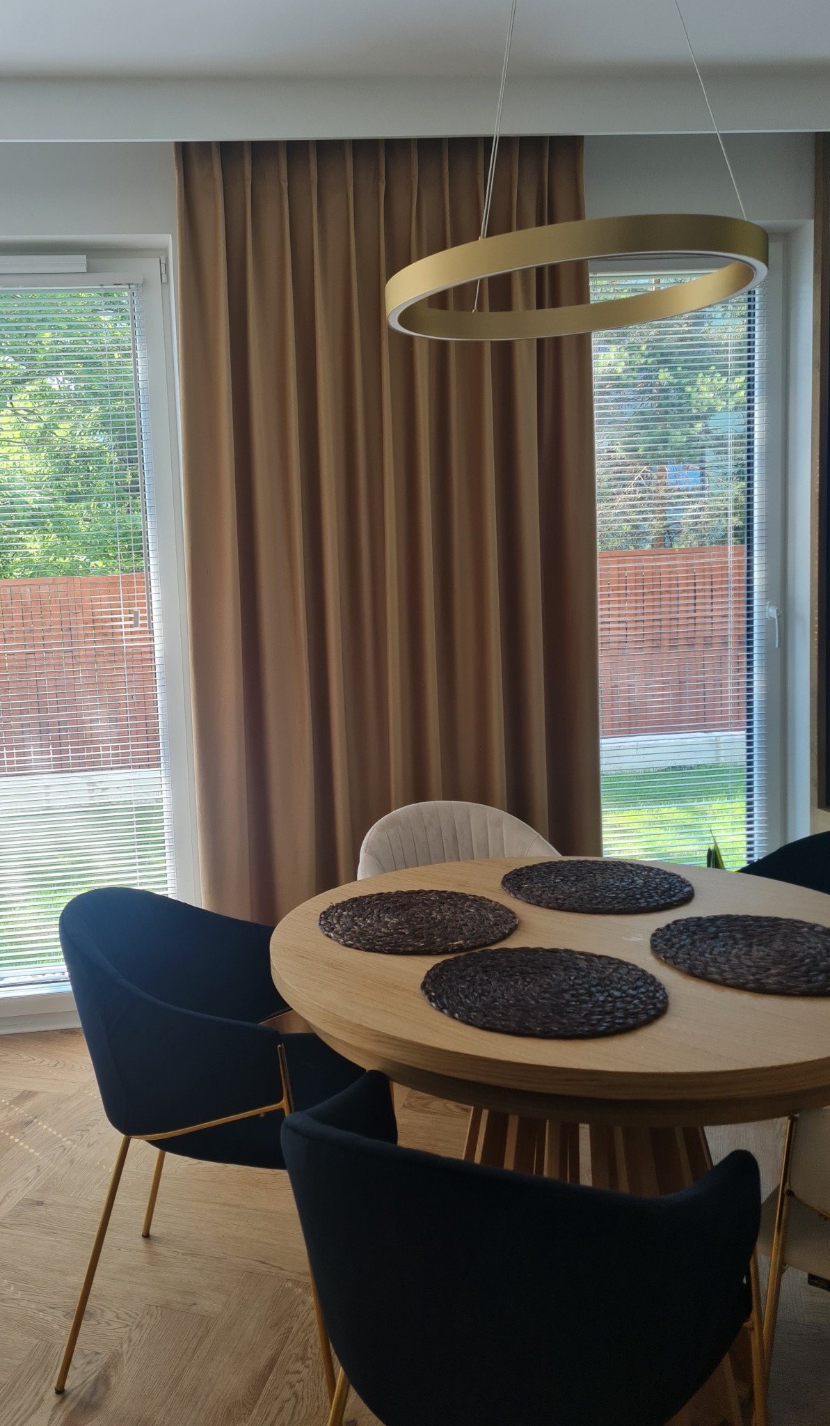 Beżowe, gładkie zasłony w nowoczesnym wnętrzu z okrągłym, drewnianym stołem, czarnymi krzesłami na złotych nogach i okrągłą lampą wiszącą.
