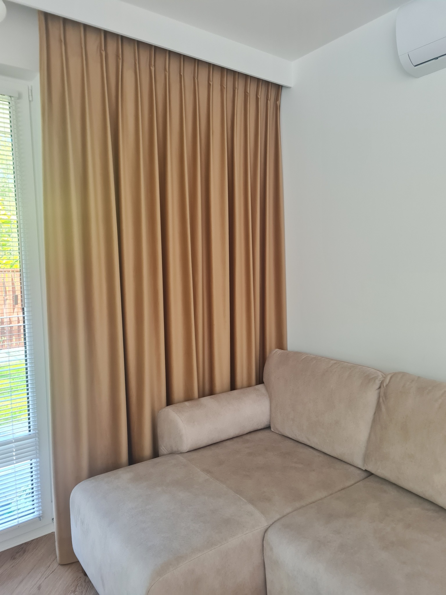 Beżowe zasłony w salonie z beżową sofą, widoczne żaluzje w oknie i klimatyzator na ścianie.