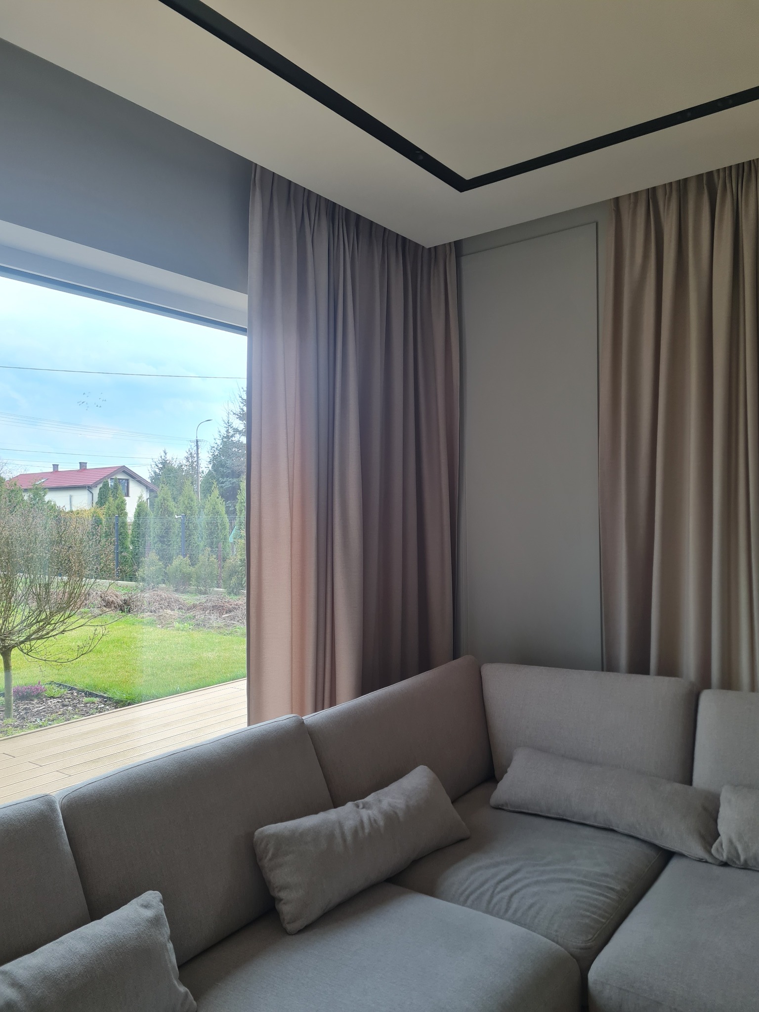 Beżowe, grube zasłony wiszące przy dużym oknie z widokiem na zielony ogród z domem; szara sofa z poduszkami w rogu pokoju.