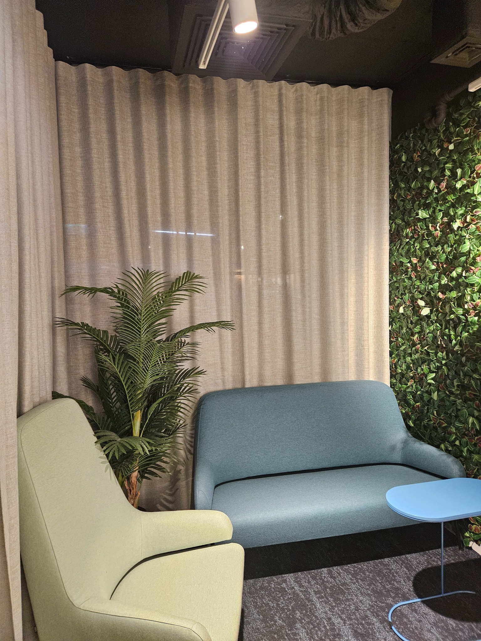 Beżowe, marszczone zasłony w biurowym wnętrzu z zieloną ścianą, jasnym fotelem, niebieską sofą i stolikiem kawowym.