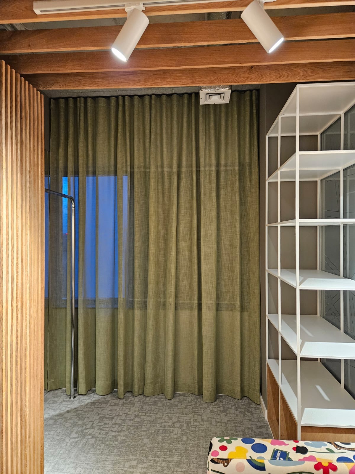 Zasłony w odcieniach zieleni, wiszące w pomieszczeniu z drewnianym sufitem i białym regałem, oświetlone punktowymi lampami.