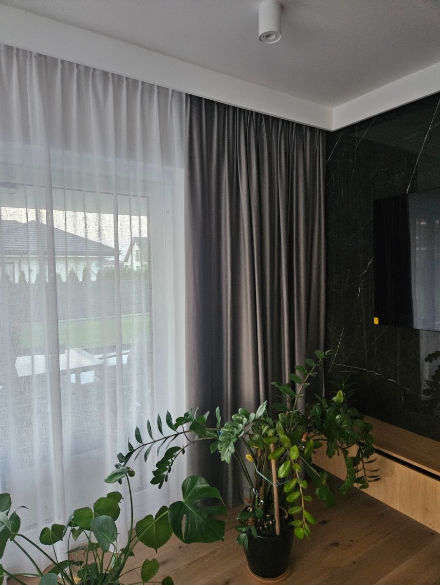 Białe i szare zasłony w oknie w Warszawie, z roślinami doniczkowymi na pierwszym planie i marmurową ścianą w tle.