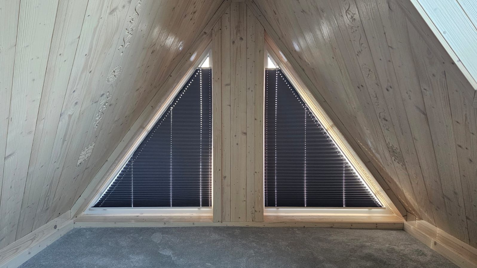 Dwa trójkątne okna na poddaszu z drewnianą boazerią i zamontowanymi plisowanymi, zaciemniającymi zasłonami w kolorze grafitowym.