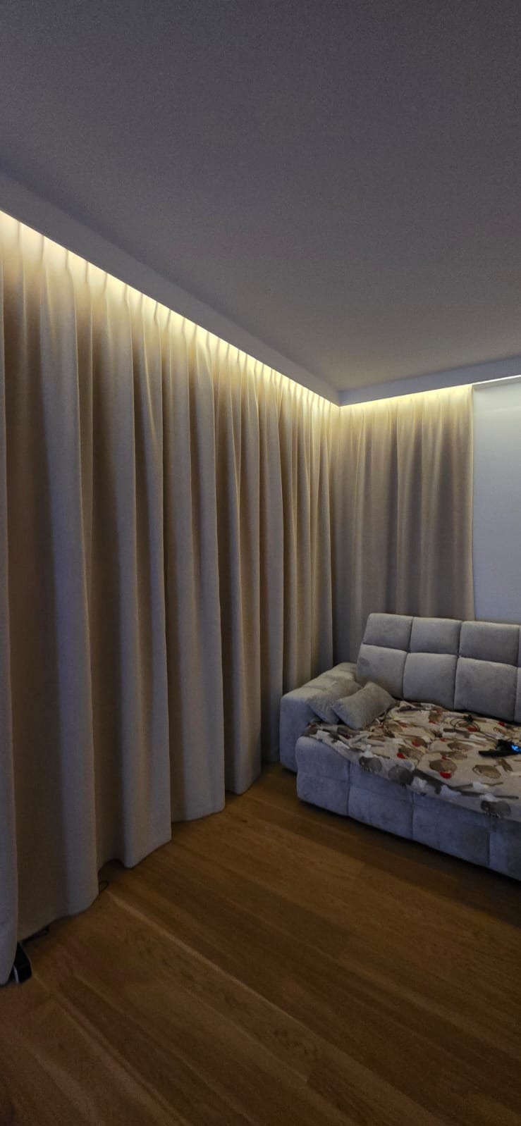Beżowe, gładkie zasłony z podświetleniem LED wzdłuż karnisza, tworzące przytulną atmosferę w pokoju w Warszawie. Widoczny fragment sofy i drewniana podłoga.