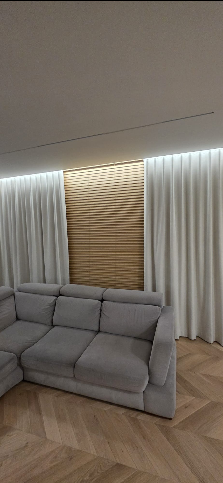 Eleganckie, długie, kremowe zasłony po bokach okna z drewnianymi żaluzjami, uzupełnione przez szarą, narożną sofę i parkiet w jodełkę.