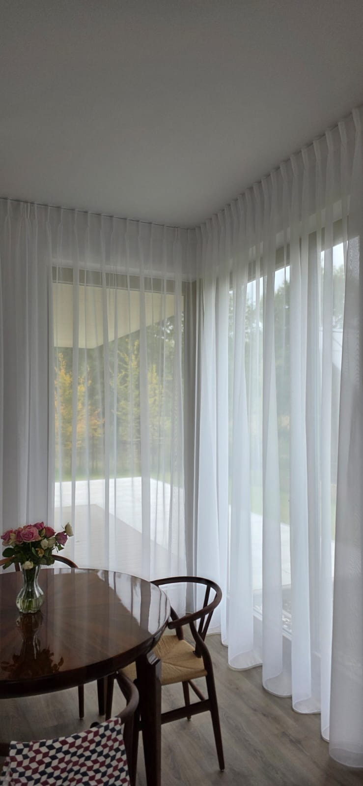 Delikatne, białe, zwiewne zasłony woalowe w jasnym wnętrzu z drewnianym stołem i bukietem róż, w tle widok na zieleń za oknem.