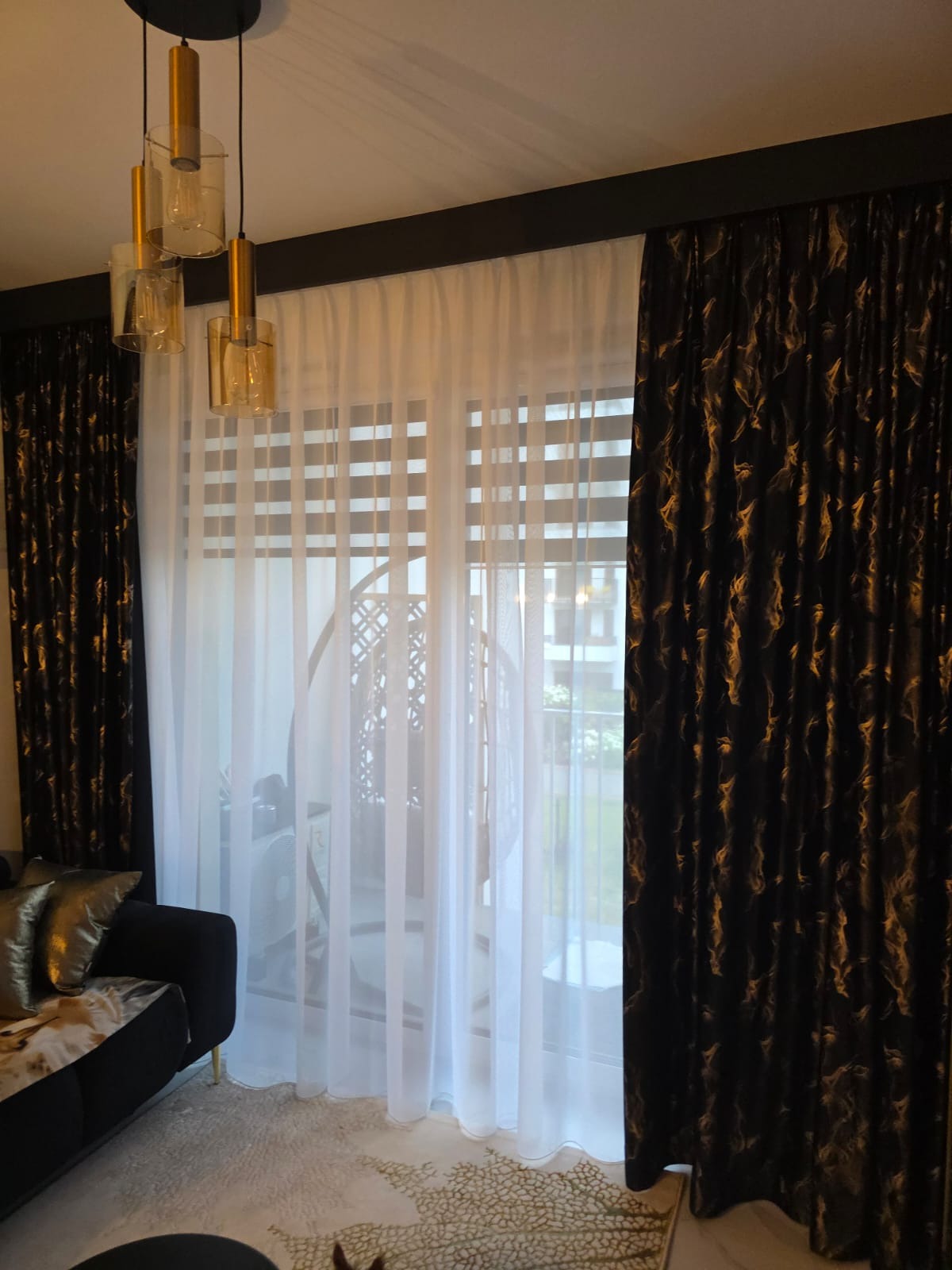 Eleganckie, długie zasłony: ciemne, wzorzyste po bokach i białe, prześwitujące na środku, w tle widoczny fotel wiszący i rolety okienne.