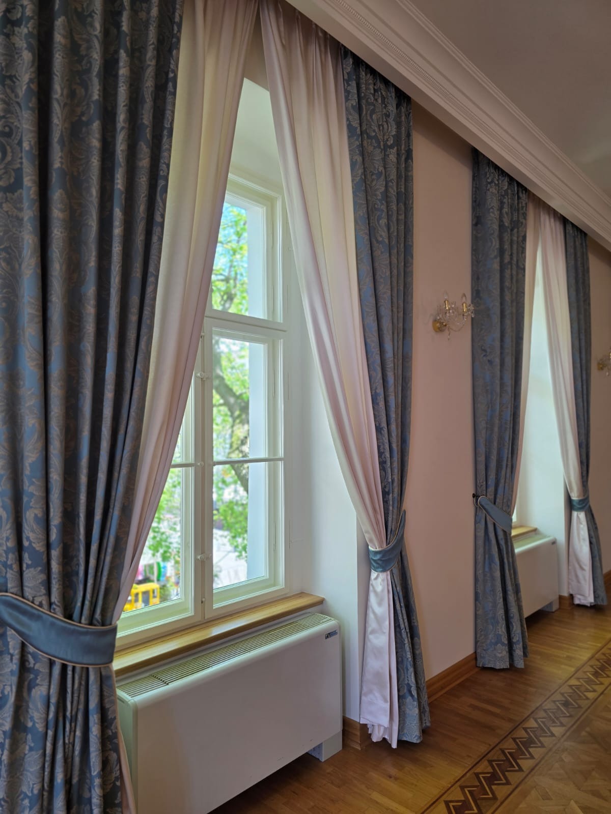 Eleganckie, dwukolorowe zasłony w klasycznym wnętrzu. Błękitne, wzorzyste zasłony spięte dekoracyjnymi taśmami, zestawione z jasnymi firanami. Widok na okno i parkiet.
