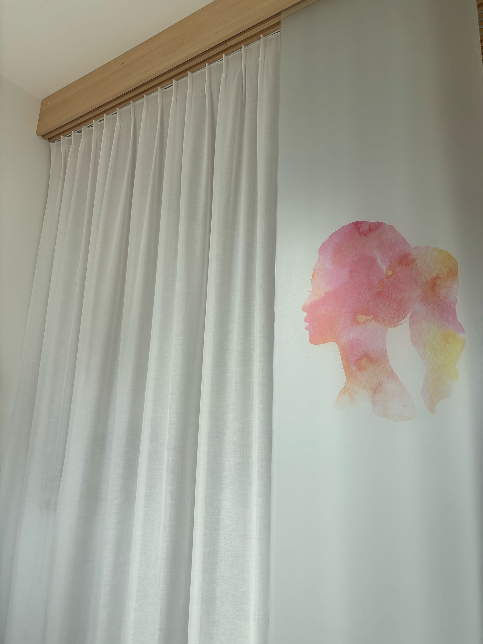 Delikatne, białe zasłony z akwarelowym motywem profilu kobiety, zawieszone na drewnianej szynie. Subtelny akcent dekoracyjny w jasnym wnętrzu.