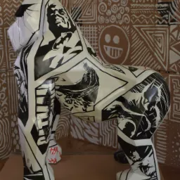 Biała figura goryla ozdobiona czarno-białymi motywami komiksowymi, w tym postaciami z Batmana, umieszczona na drewnianej palecie, na tle ściany z geometrycznymi wzorami.