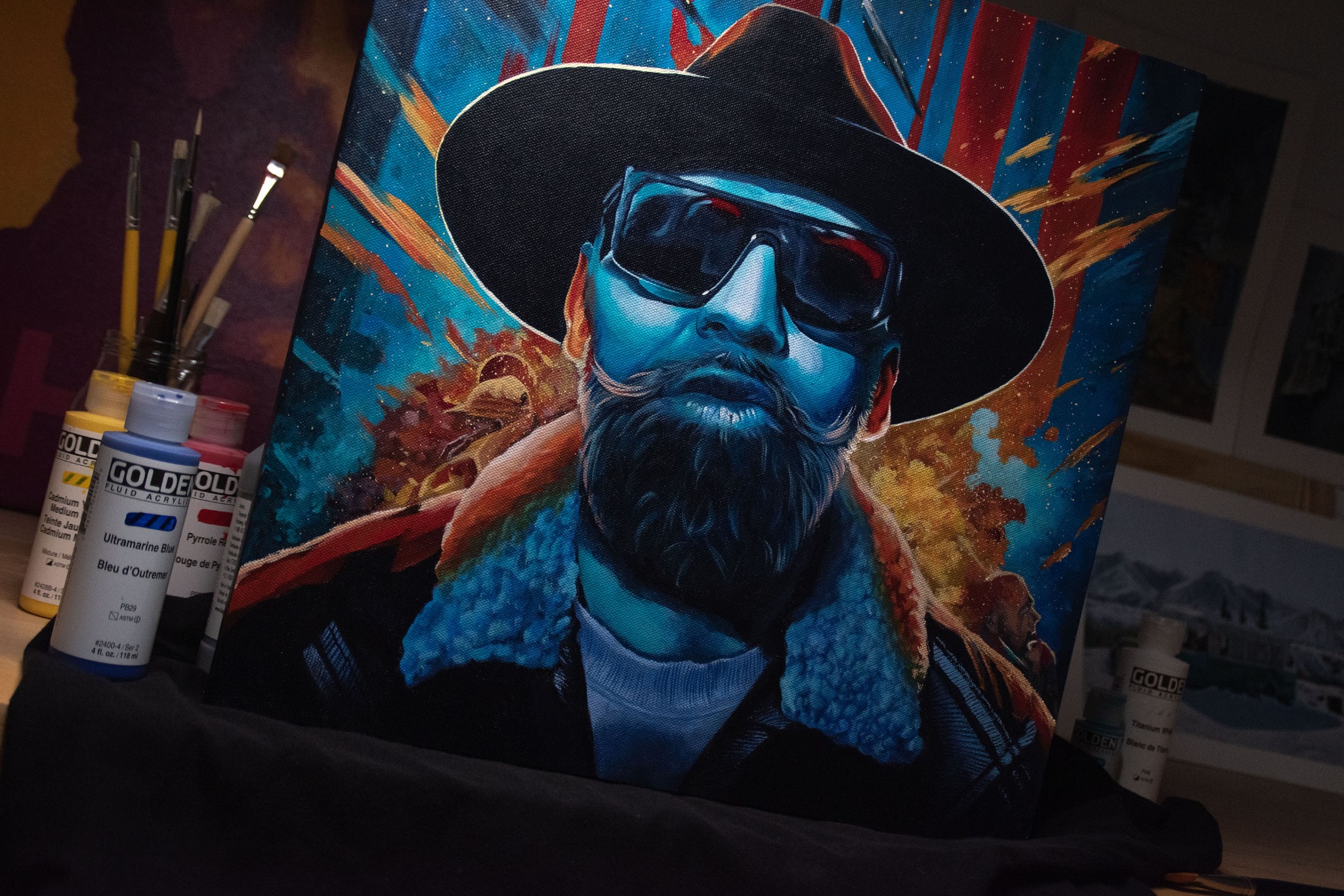 Portret mężczyzny z brodą i wąsami w kapeluszu i okularach przeciwsłonecznych, namalowany w stylu pop-art na płótnie, obok farb akrylowych i pędzli.