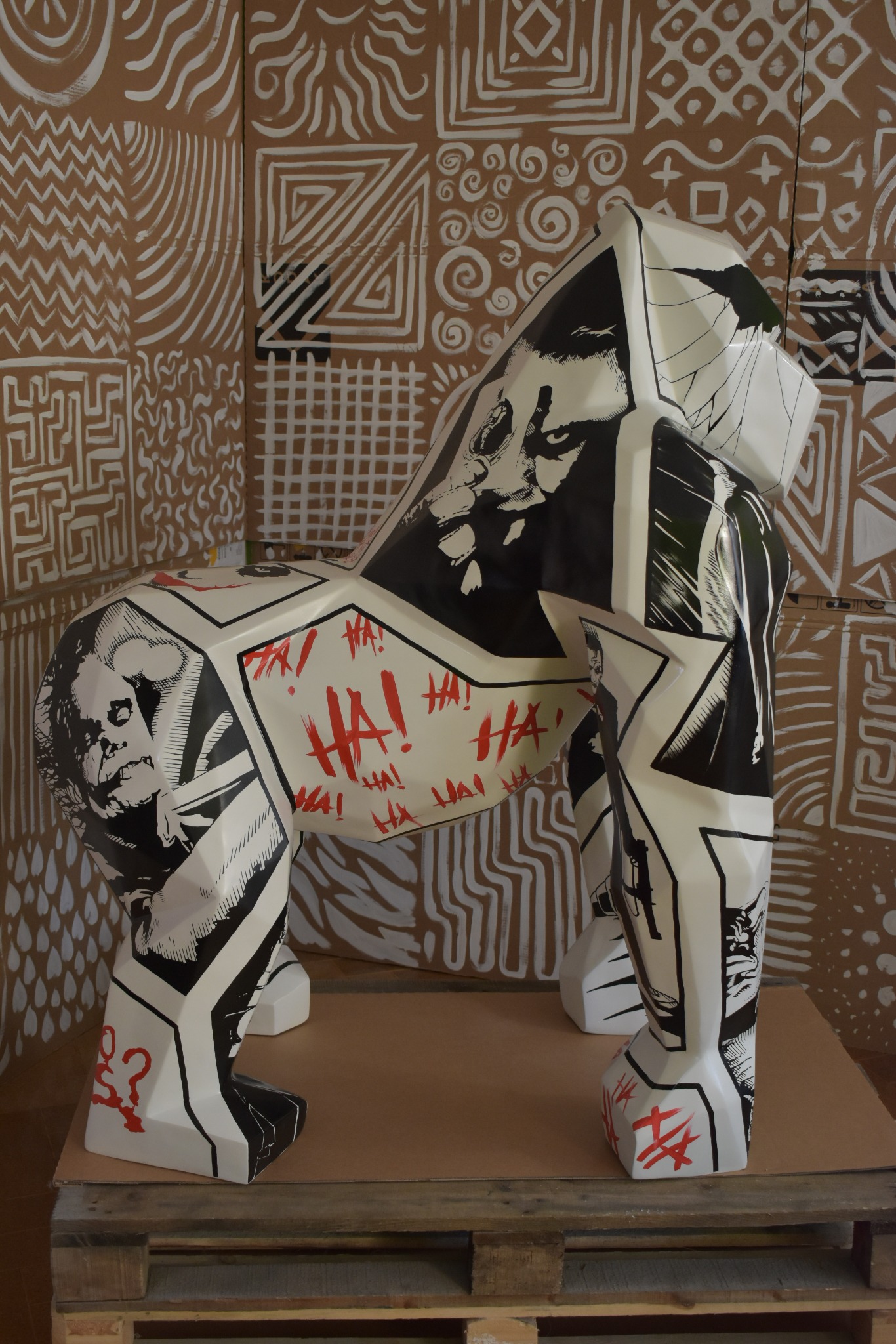 Rzeźba goryla z geometrycznymi wzorami i motywami Jokera, umieszczona na palecie na tle kartonowej ściany z białymi, abstrakcyjnymi malunkami.