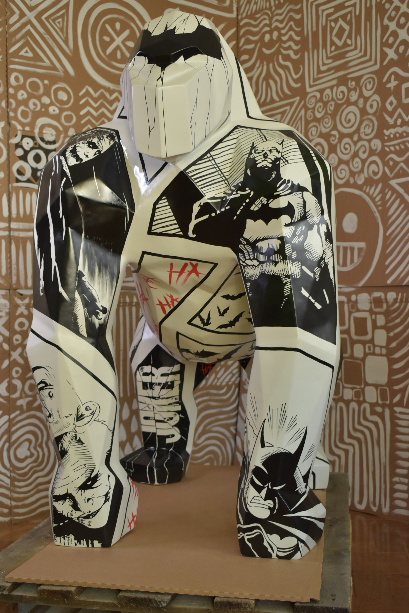 Biała, geometryczna rzeźba goryla z czarno-białymi grafikami Batmana i Jokera, ustawiona na drewnianej podstawie na tle ściany z plemiennymi wzorami.