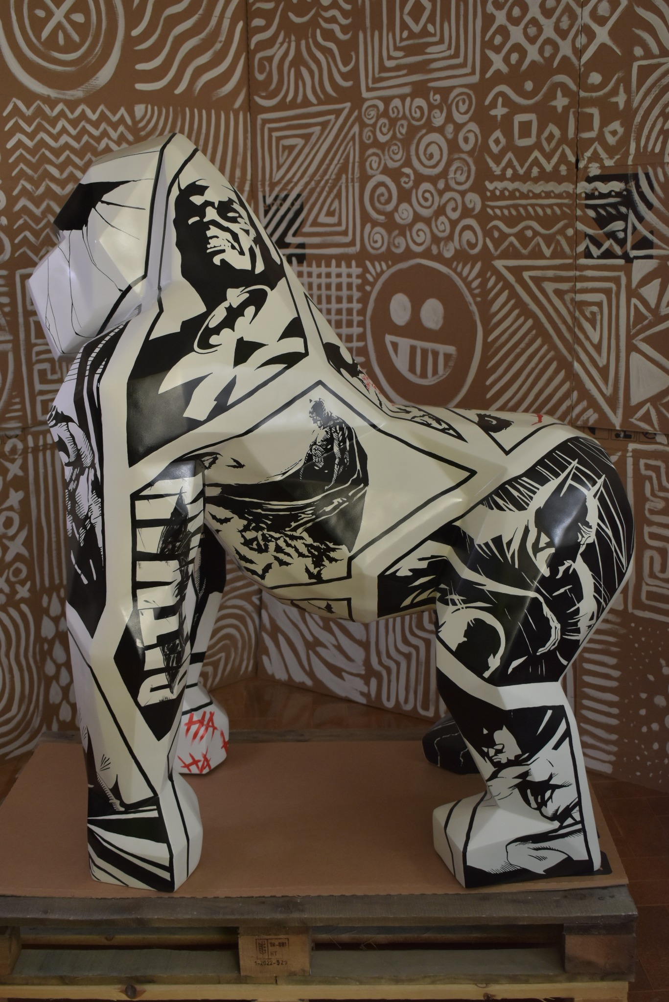 Biała figura goryla ozdobiona czarno-białymi motywami komiksowymi, w tym postaciami z Batmana, umieszczona na drewnianej palecie, na tle ściany z geometrycznymi wzorami.