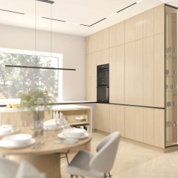 KB Interior Design Karol Budziszewski - Jasna kuchnia z drewnianymi frontami, okrągłym stołem i krzesłami. Minimalistyczne oświetlenie i zabudowa piekarnika. Stonowana kolorystyka wnętrza, widok na zieleń za oknem.