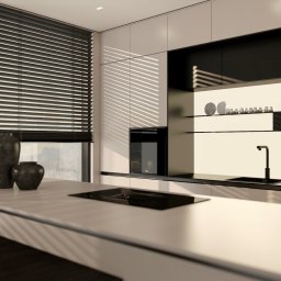 KB Interior Design Karol Budziszewski - Minimalistyczna kuchnia w bieli i czerni z żaluzjami poziomymi, oświetlona promieniami słońca. Na blacie dekoracyjne wazony, w tle zabudowa kuchenna z piekarnikiem i półkami.