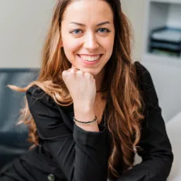 Uśmiechnięta kobieta w czarnej marynarce, opierająca brodę na pięści, z widocznym złotym zegarkiem na nadgarstku i bransoletką z czarnych i przezroczystych koralików, pozująca w jasnym biurze.