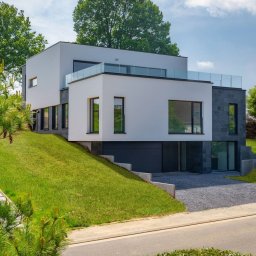 AW CENTRUM BUDOWNICTWO-REMONTY-INSTALACJE - Nowoczesny dom z białymi ścianami i elementami z szarego kamienia, z płaskim dachem i szklaną balustradą na tarasie. Uwagę przykuwa garaż wkomponowany w zbocze.