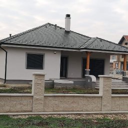 AW CENTRUM BUDOWNICTWO-REMONTY-INSTALACJE - Parterowy dom z ciemną dachówką i jasną elewacją, widoczny z przodu, z niskim murem z bloków. Elementy drewniane pod dachem. Szare okiennice i kominy.