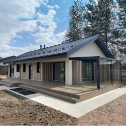 AW CENTRUM BUDOWNICTWO-REMONTY-INSTALACJE - Nowoczesny dom parterowy z drewnianą elewacją, szarą blachodachówką i przeszkloną werandą w słoneczny dzień. Widoczny taras i zadbany teren wokół budynku.