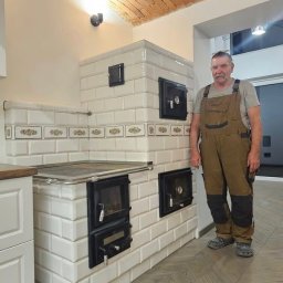 AW CENTRUM BUDOWNICTWO-REMONTY-INSTALACJE - Biały, kaflowy piec kuchenny z czarnymi drzwiczkami, wykończony dekoracyjnym pasem, na tle ceglanego sklepienia. Obok stoi mężczyzna w kombinezonie.