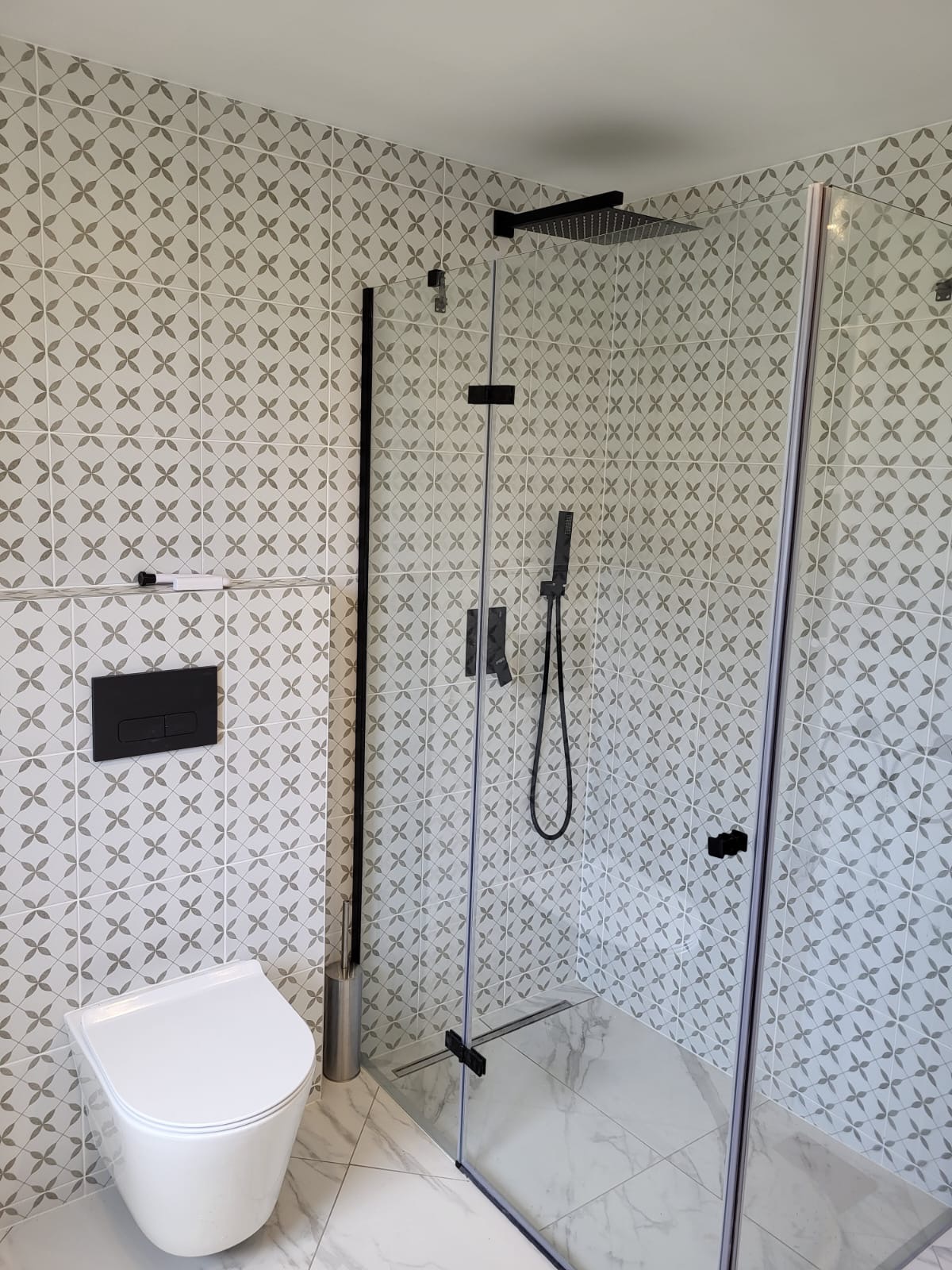 Nowoczesna łazienka z kabiną prysznicową typu walk-in, białą wiszącą toaletą i wzorzystymi szaro-białymi płytkami na ścianach i podłodze imitującymi marmur.