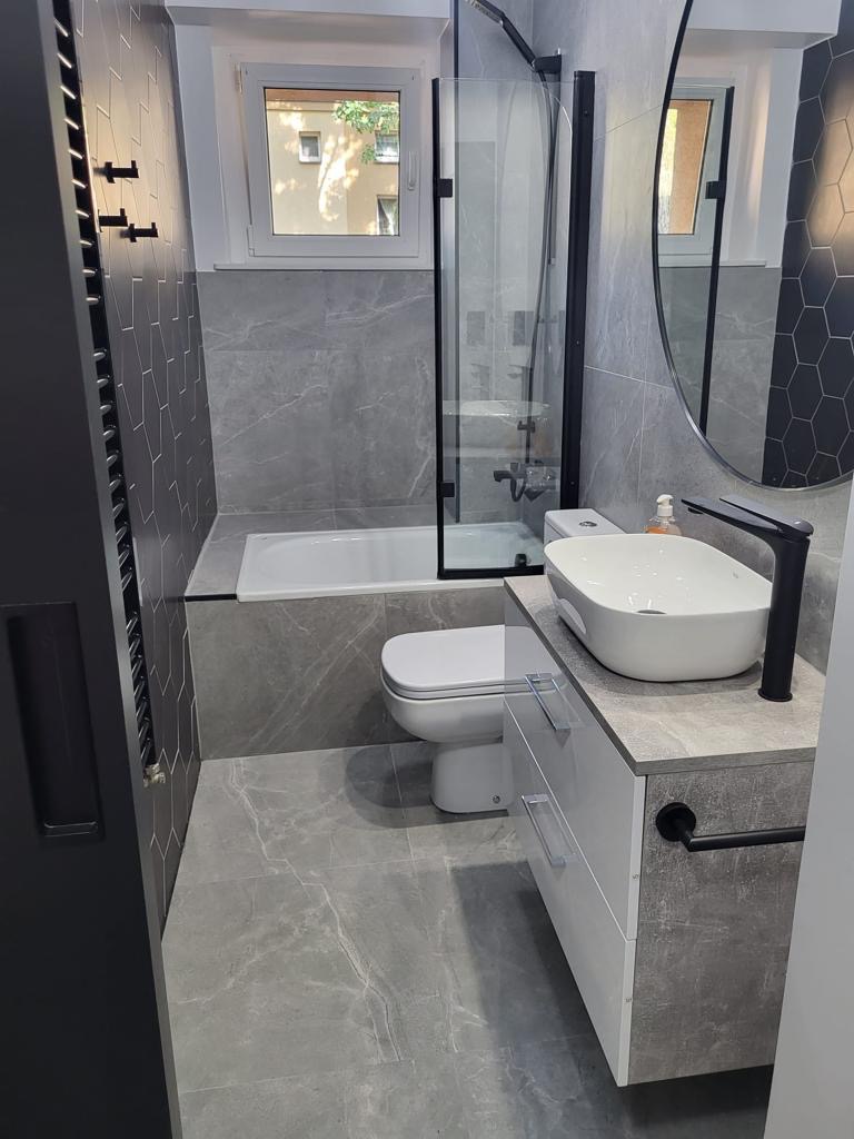 Nowoczesna łazienka w odcieniach szarości z białą armaturą, kabiną prysznicową z czarnymi ramami, okrągłym lustrem i grzejnikiem drabinkowym, widok na fragment okna z zielenią na zewnątrz.