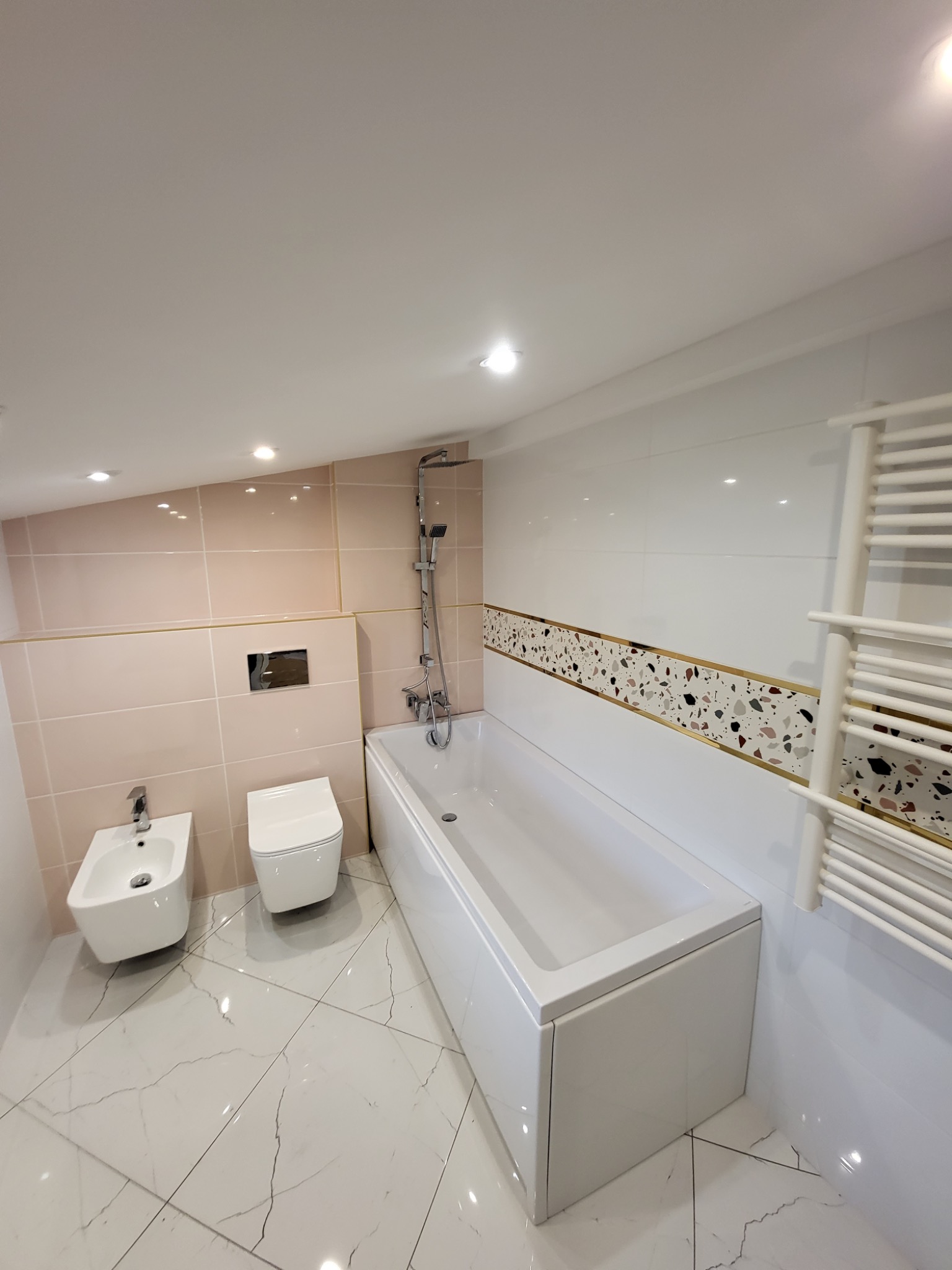 Nowoczesna łazienka z białą wanną, bidetem i toaletą, wykończona beżowymi i białymi płytkami z dekoracyjnym pasem mozaiki w odcieniach beżu, brązu i czerni, ogrzewanie drabinkowe.