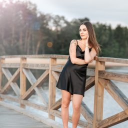 Agart - Portret kobiety w czarnej sukience, opierającej się o drewnianą barierkę mostu z widokiem na jezioro i las w tle, z tatuażem na ramieniu i złotym zegarkiem.
