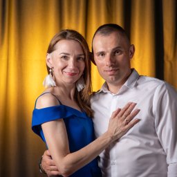 Agart - Para w objęciach na tle złotych zasłon, kobieta w kobaltowej sukience i dużych białych kolczykach, mężczyzna w białej koszuli, widoczne obrączki.