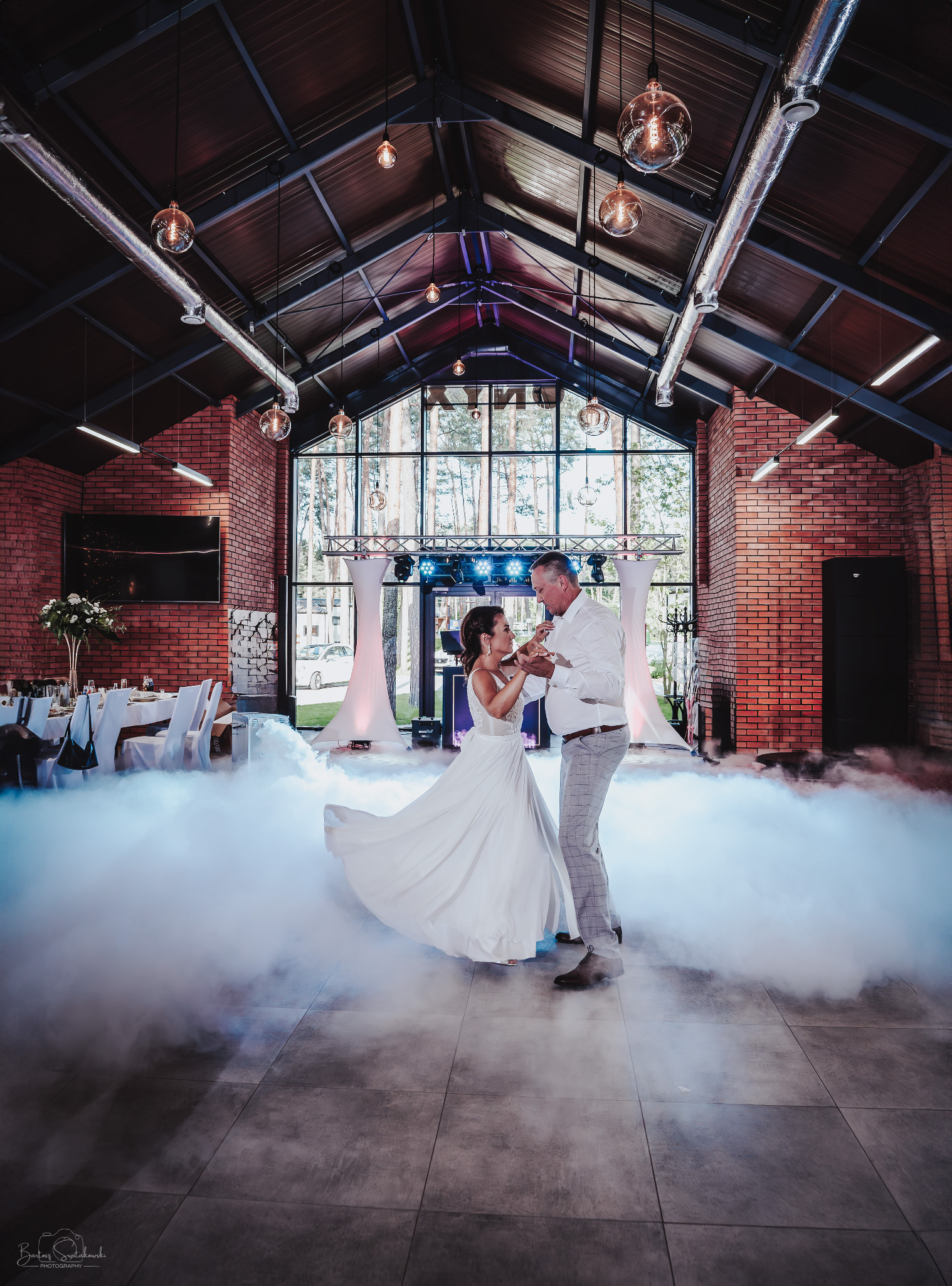 Para młoda tańczy pierwszy taniec w sali z ceglanymi ścianami, oświetloną dekoracyjnymi żarówkami i wypełnioną sztuczną mgłą.