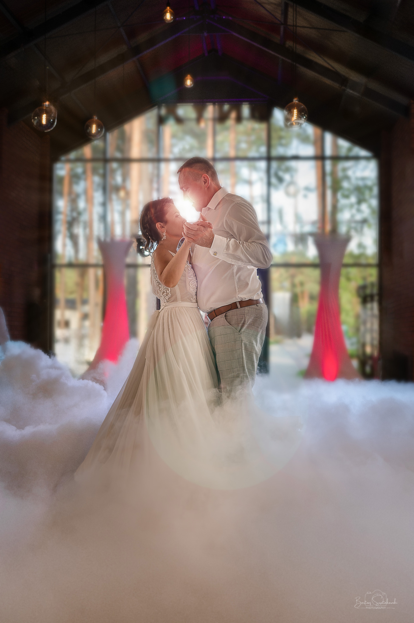 Para młoda tańczy pierwszy taniec w dymie na tle wysokich okien w przestronnej sali z żarówkami Edisona zwisającymi z sufitu.