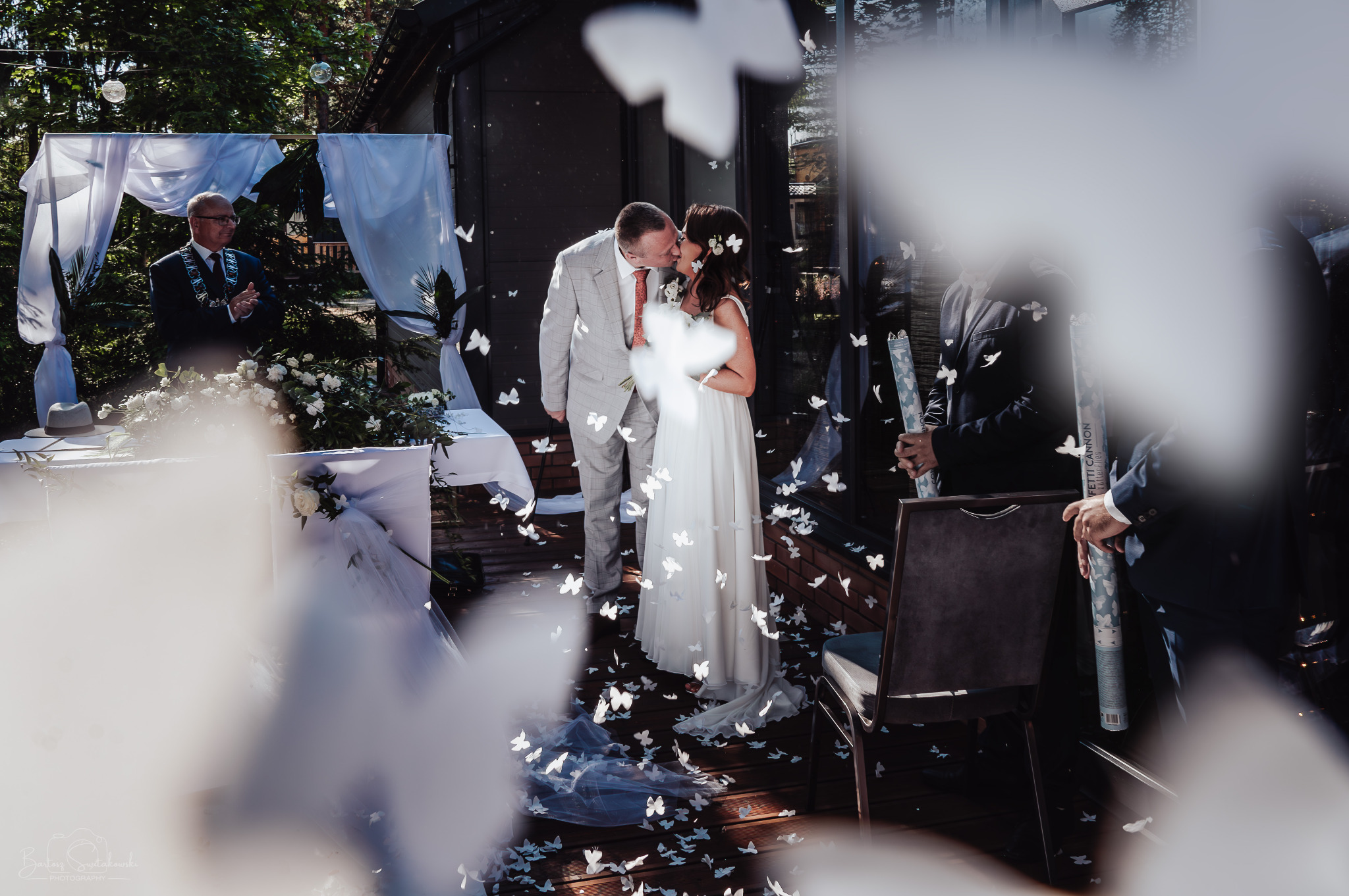 Pocałunek nowożeńców w plenerze, zasypanych konfetti w kształcie motyli, w tle dekoracja ślubna i osoba udzielająca ślubu.