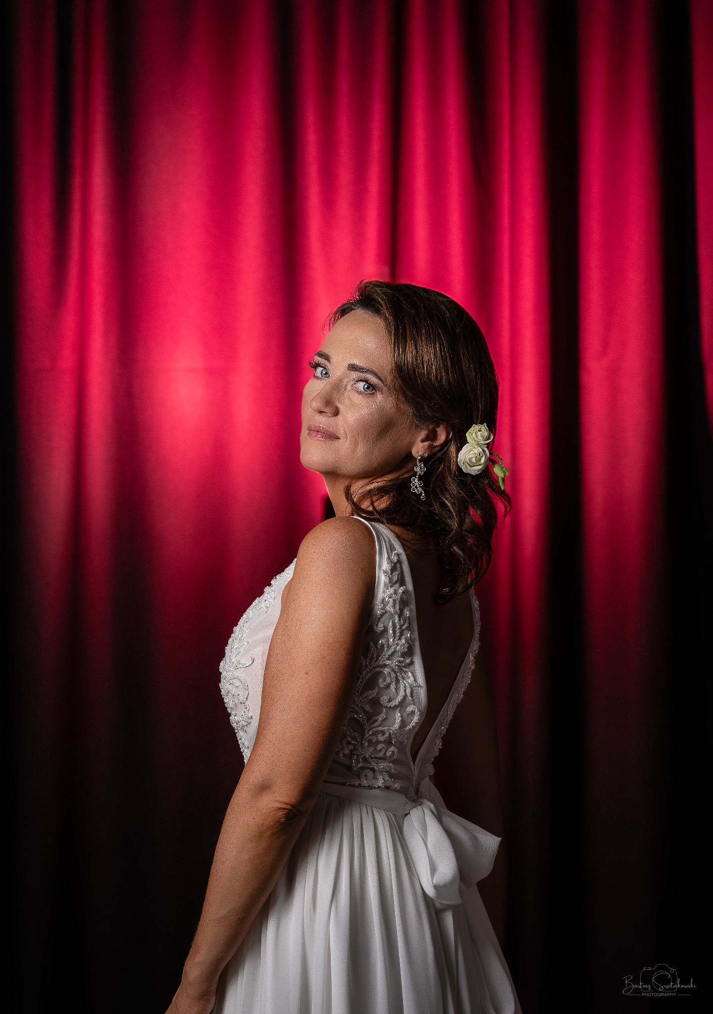 Panna młoda w białej sukni z odkrytymi plecami, z kwiatami we włosach, spogląda przez ramię na tle czerwonej zasłony.