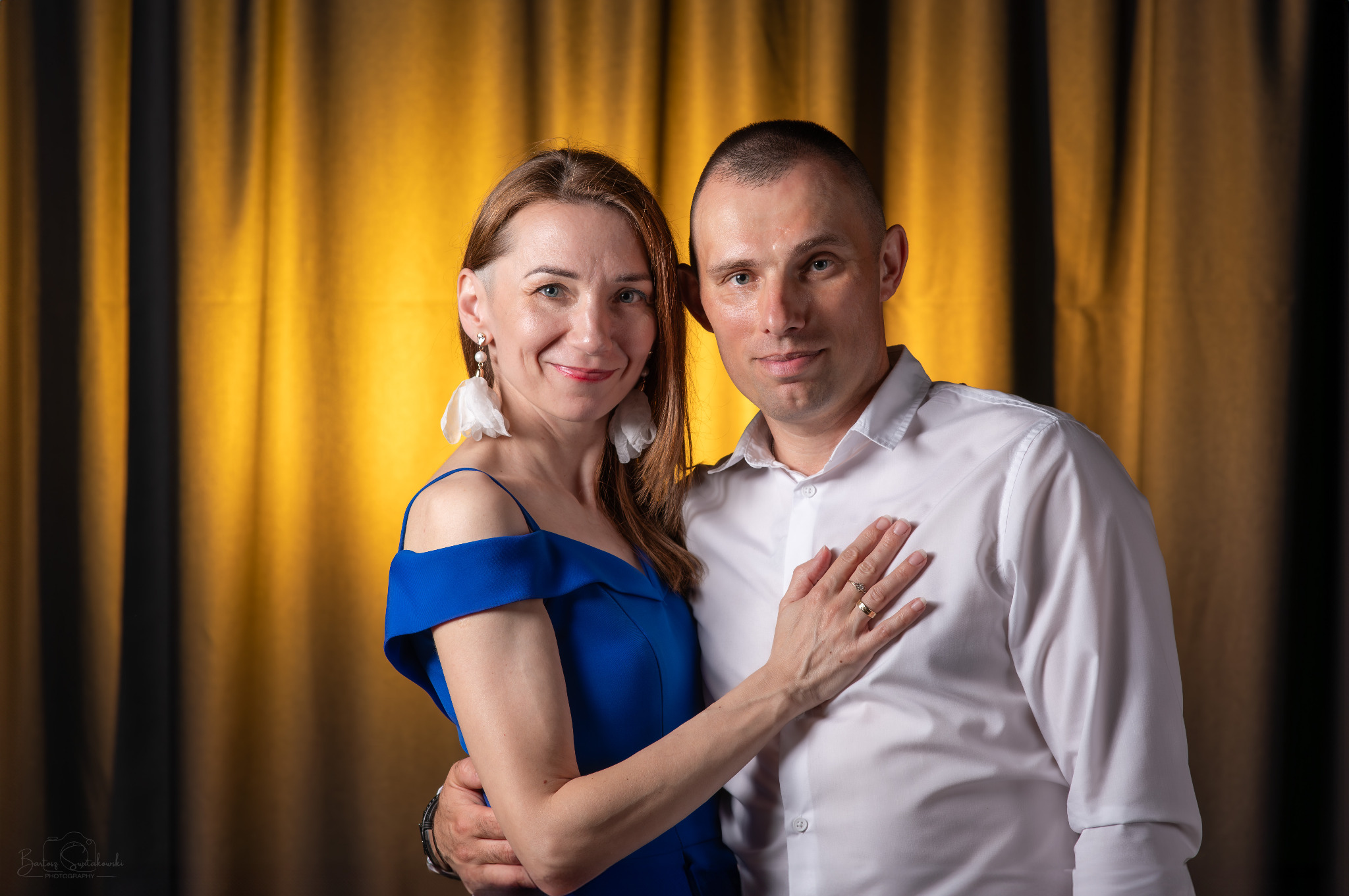 Para w objęciach na tle złotych zasłon, kobieta w kobaltowej sukience i dużych białych kolczykach, mężczyzna w białej koszuli, widoczne obrączki.