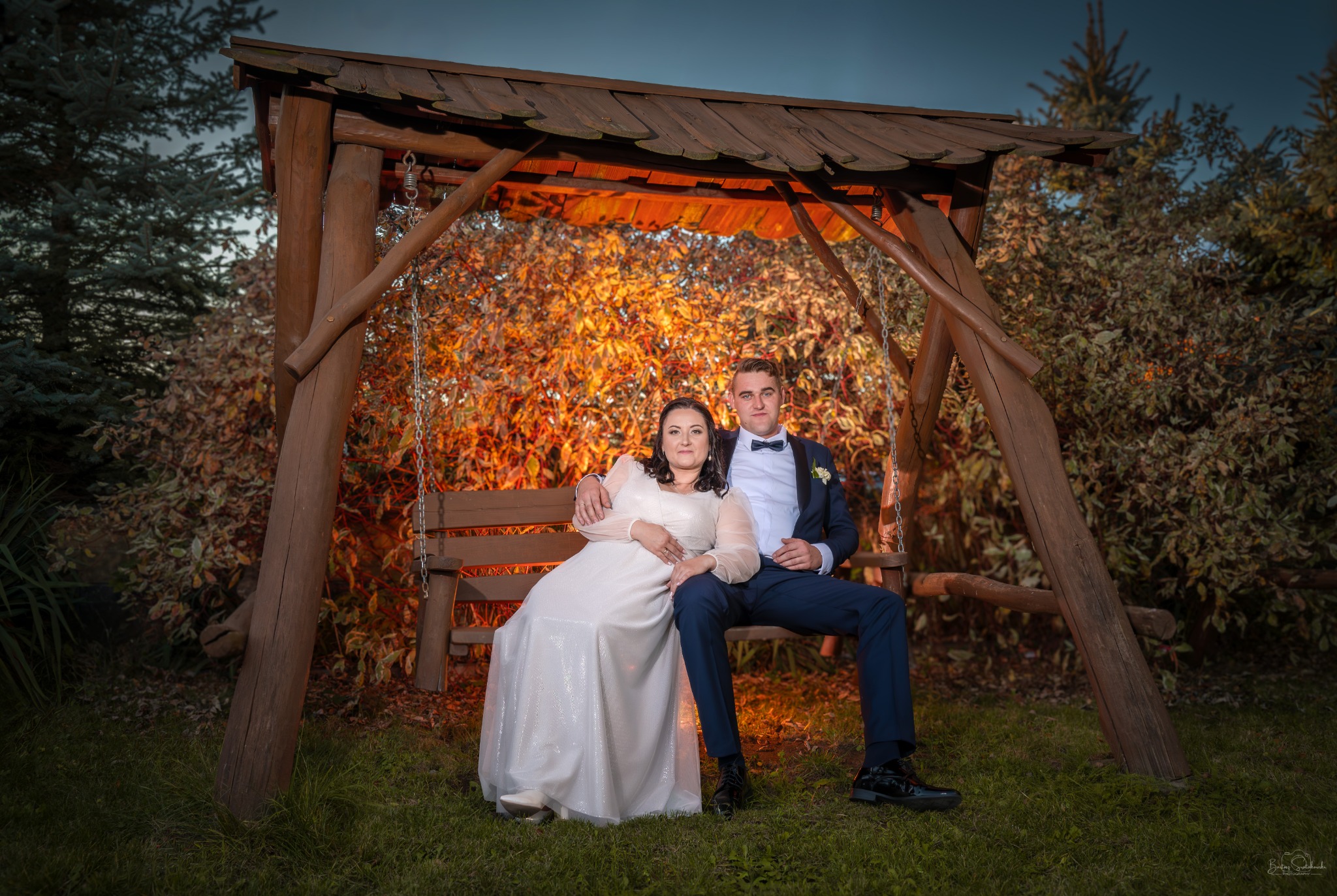 Para młoda siedząca na drewnianej huśtawce w ogrodzie, panna młoda w białej sukni, pan młody w granatowym garniturze z muszką, jesienne liście w tle.