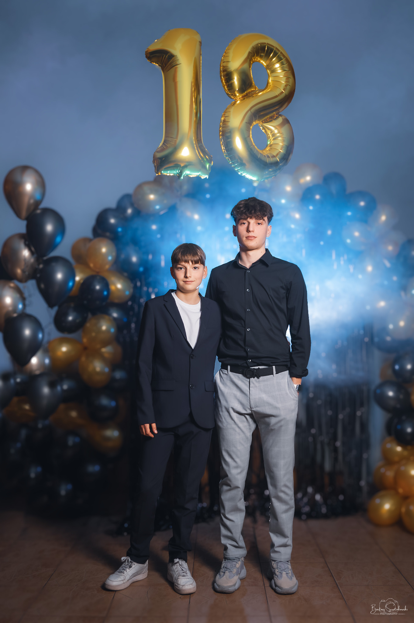 Portret dwojga nastolatków na tle z balonów i cyfr '18', chłopak w czarnej koszuli i szarych spodniach, dziewczyna w garniturze, oboje w sportowych butach.