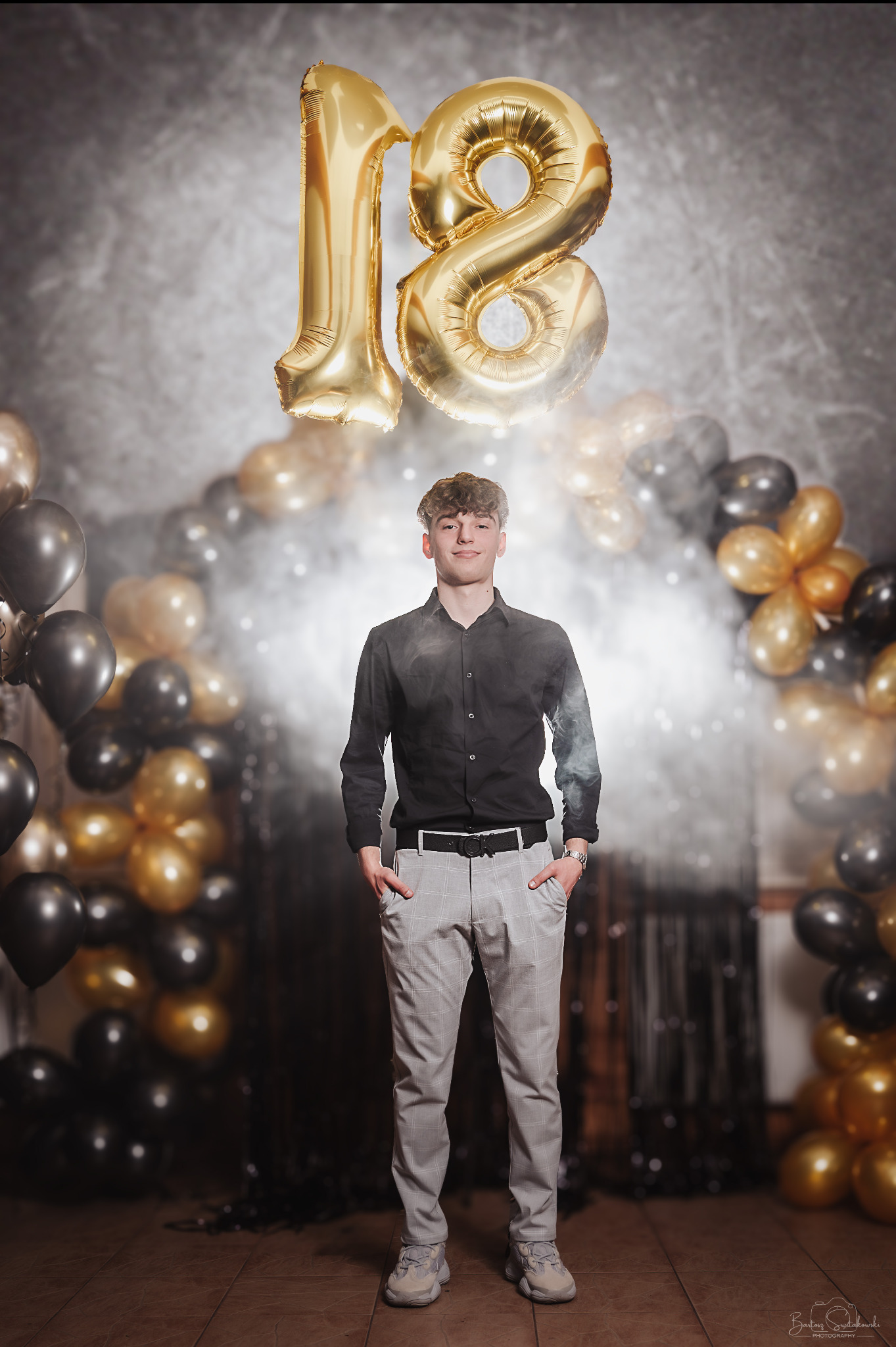Młody mężczyzna pozuje na tle dekoracji z czarno-złotych balonów i balonów foliowych w kształcie liczby 18, ubrany w czarną koszulę i kraciaste spodnie.