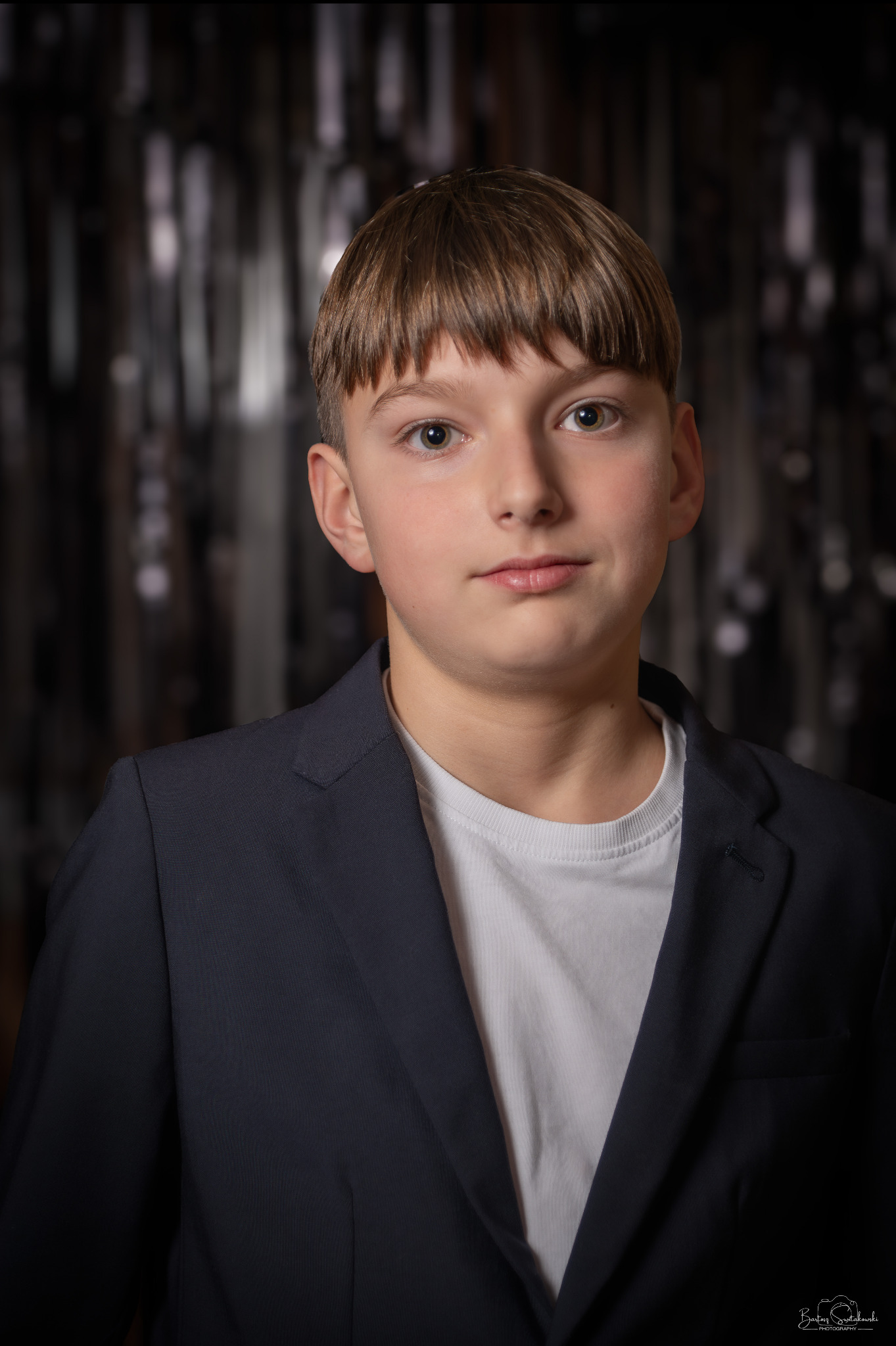 Portret młodego chłopca w garniturze na tle błyszczącej, abstrakcyjnej dekoracji.