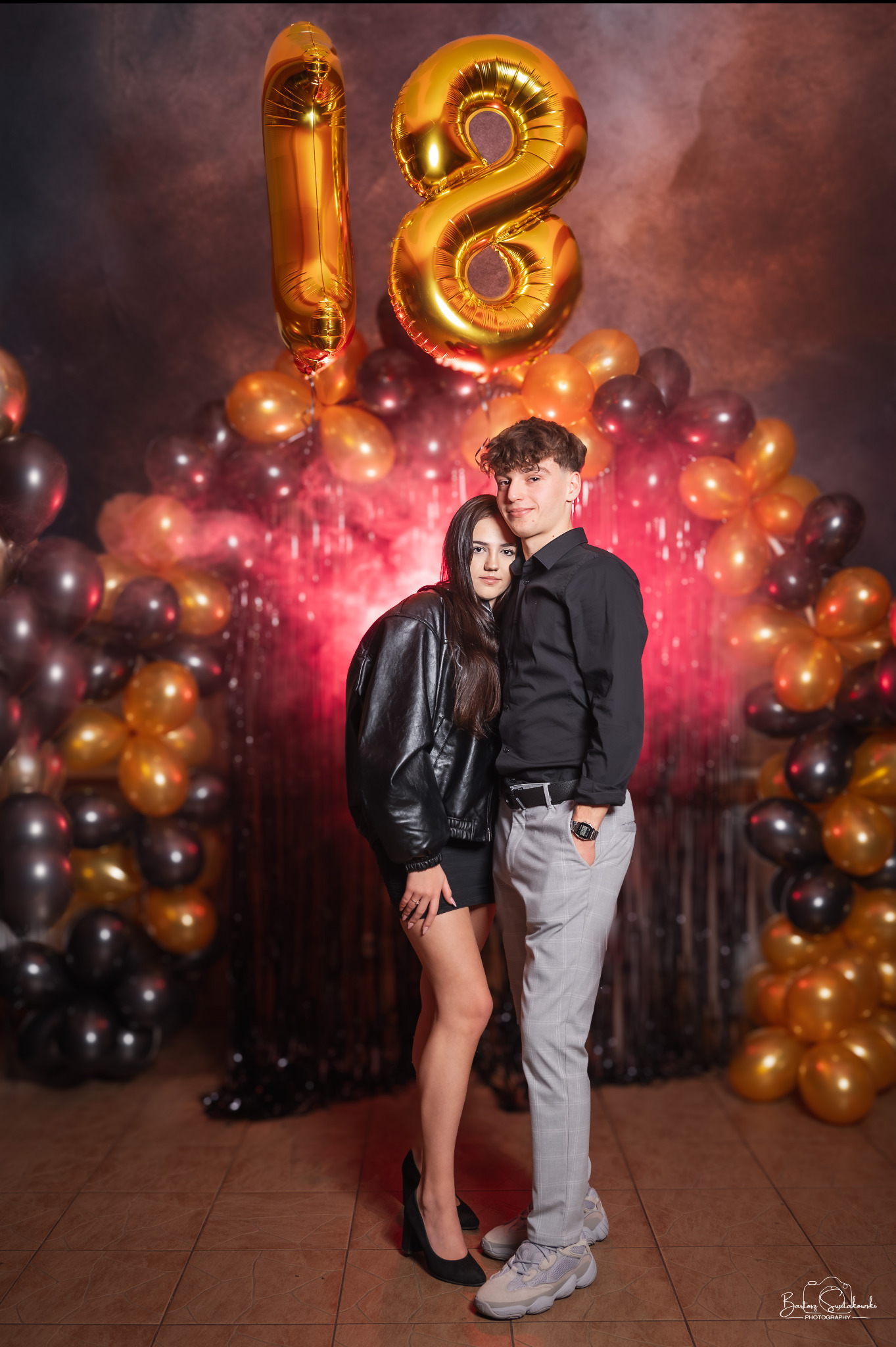 Para nastolatków pozuje na tle dekoracji urodzinowych z balonami w kształcie cyfry 18, ona w czarnej skórzanej kurtce i szpilkach, on w czarnej koszuli i szarych spodniach w kratę, z ręką w kieszeni.