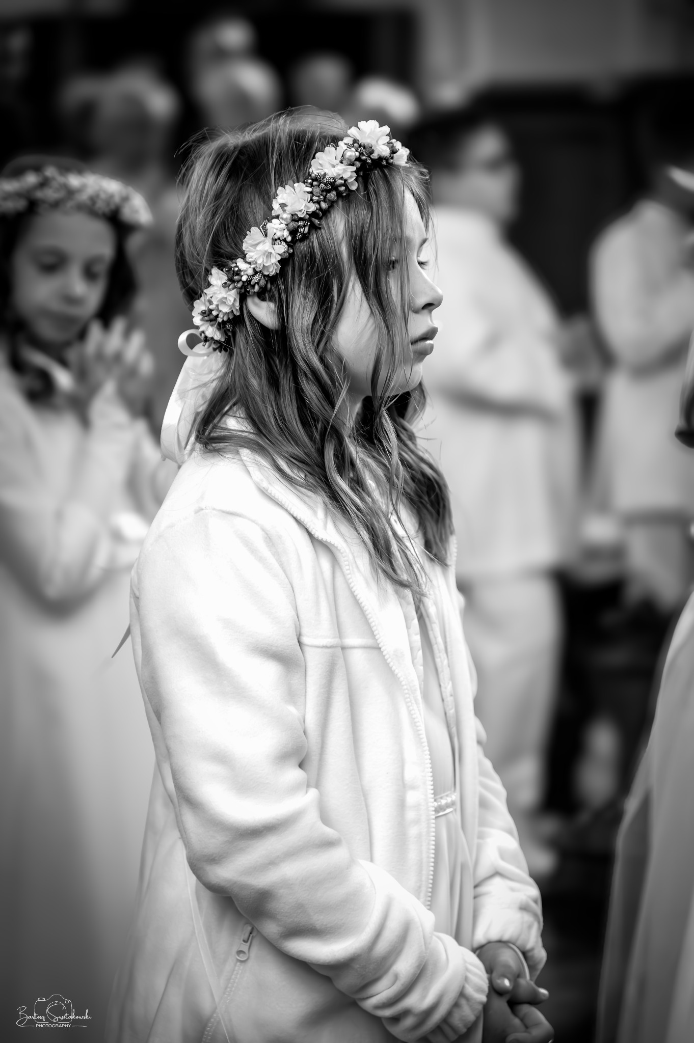 Czarno-białe zdjęcie dziewczynki z wiankiem na głowie, profil, zamyślony wyraz twarzy, w tle inne dzieci ubrane na biało, prawdopodobnie uroczystość.