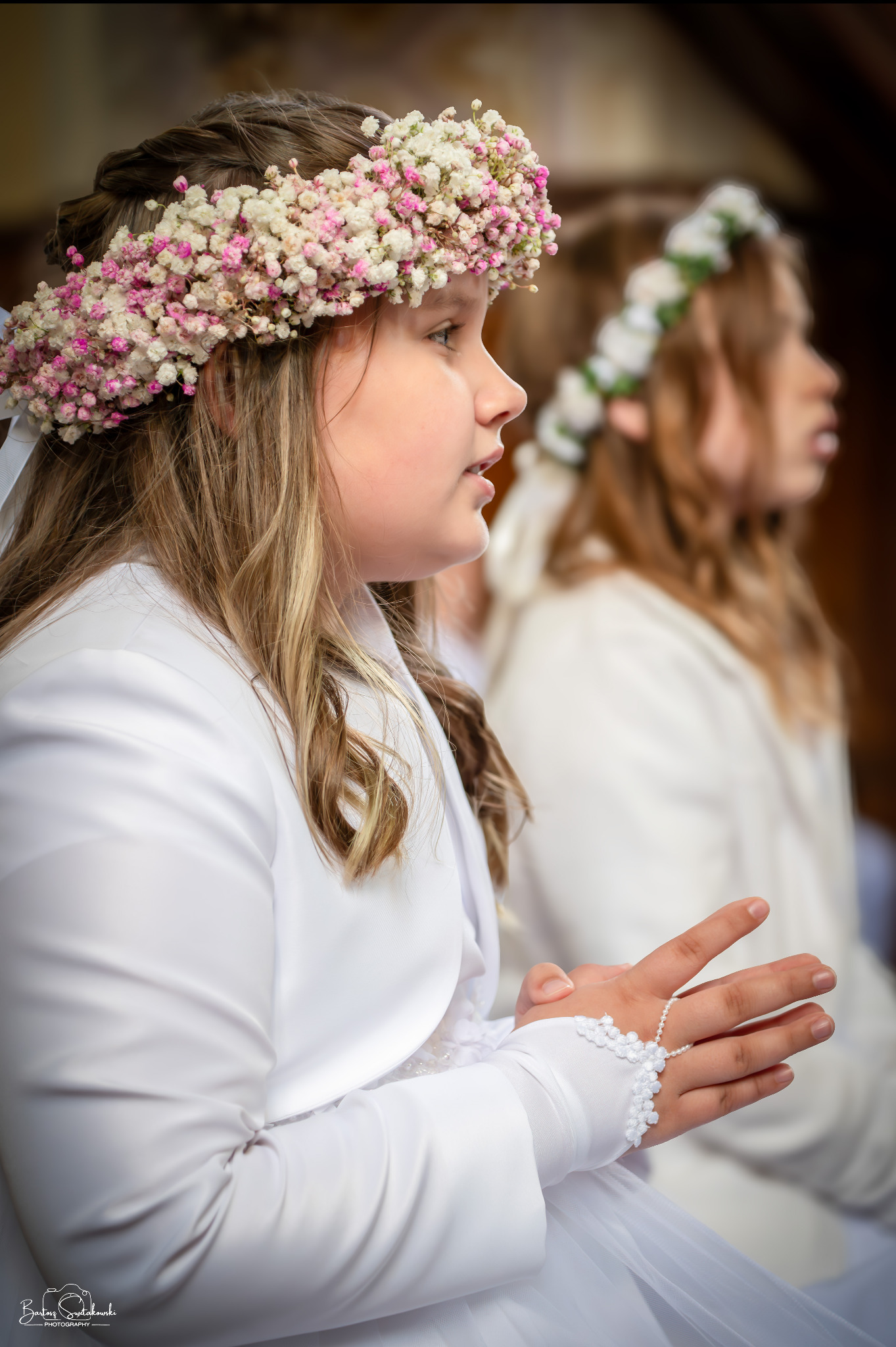 Dziewczynka w białym stroju komunijnym i wianku z gipsówki na głowie, profil, złożone dłonie, w tle inne dzieci w strojach komunijnych. Delikatne światło.