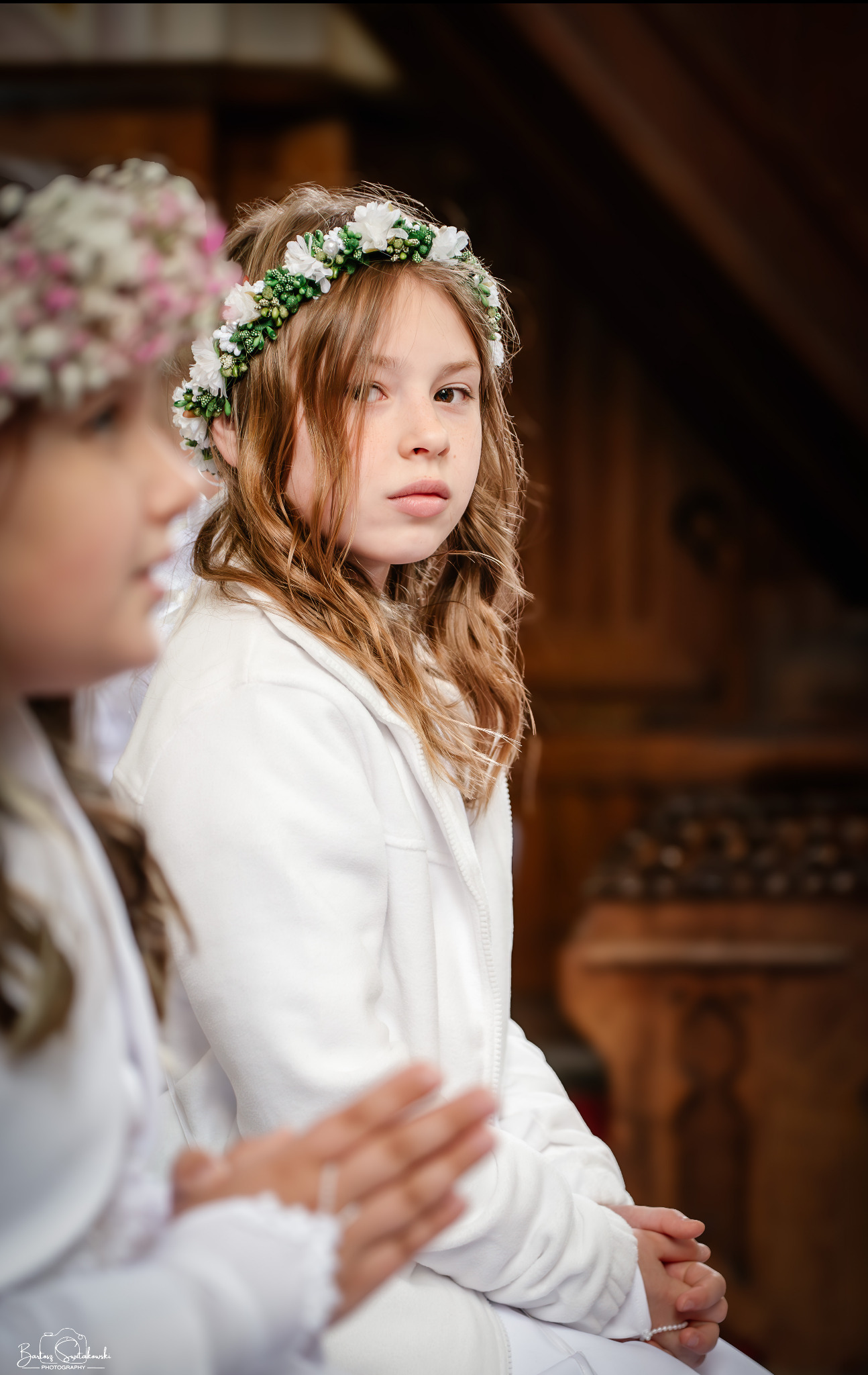 Portret dziewczynki w wianku z białych kwiatów podczas Pierwszej Komunii Świętej. Ujęcie z kościoła, w tle drewniane elementy wyposażenia. Spokojny wyraz twarzy.