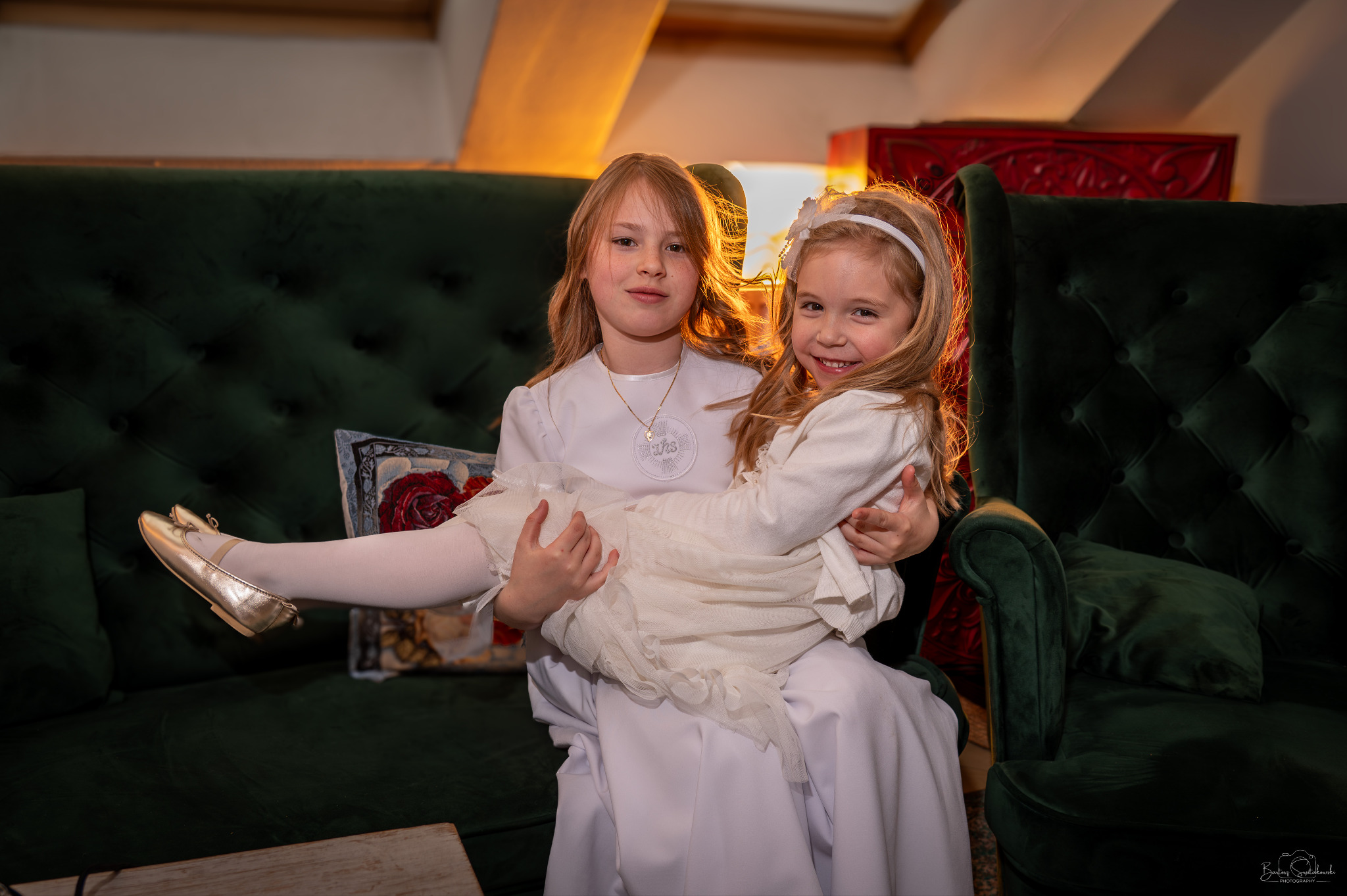 Dwie dziewczynki w białych strojach komunijnych siedzą na zielonej, pikowanej sofie. Jedna trzyma drugą na kolanach, widoczne złote baleriny i uśmiechnięte twarze.