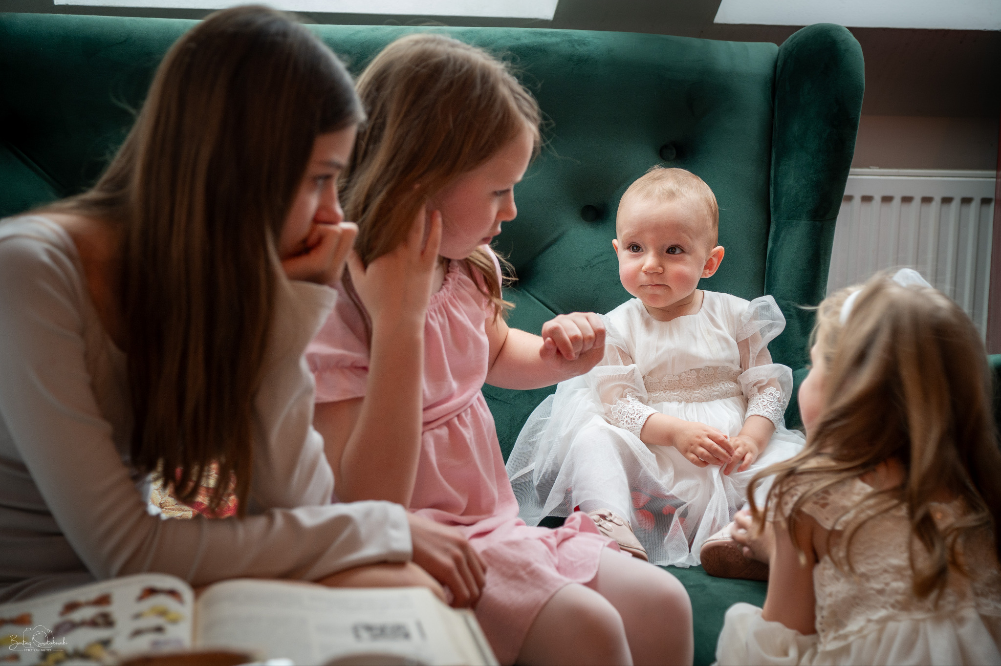 Trzy dziewczynki w sukienkach siedzą na zielonej sofie. Jedna w białej sukience siedzi, a dwie w różowych sukienkach siedzą obok. Jedna z nich czyta książkę.