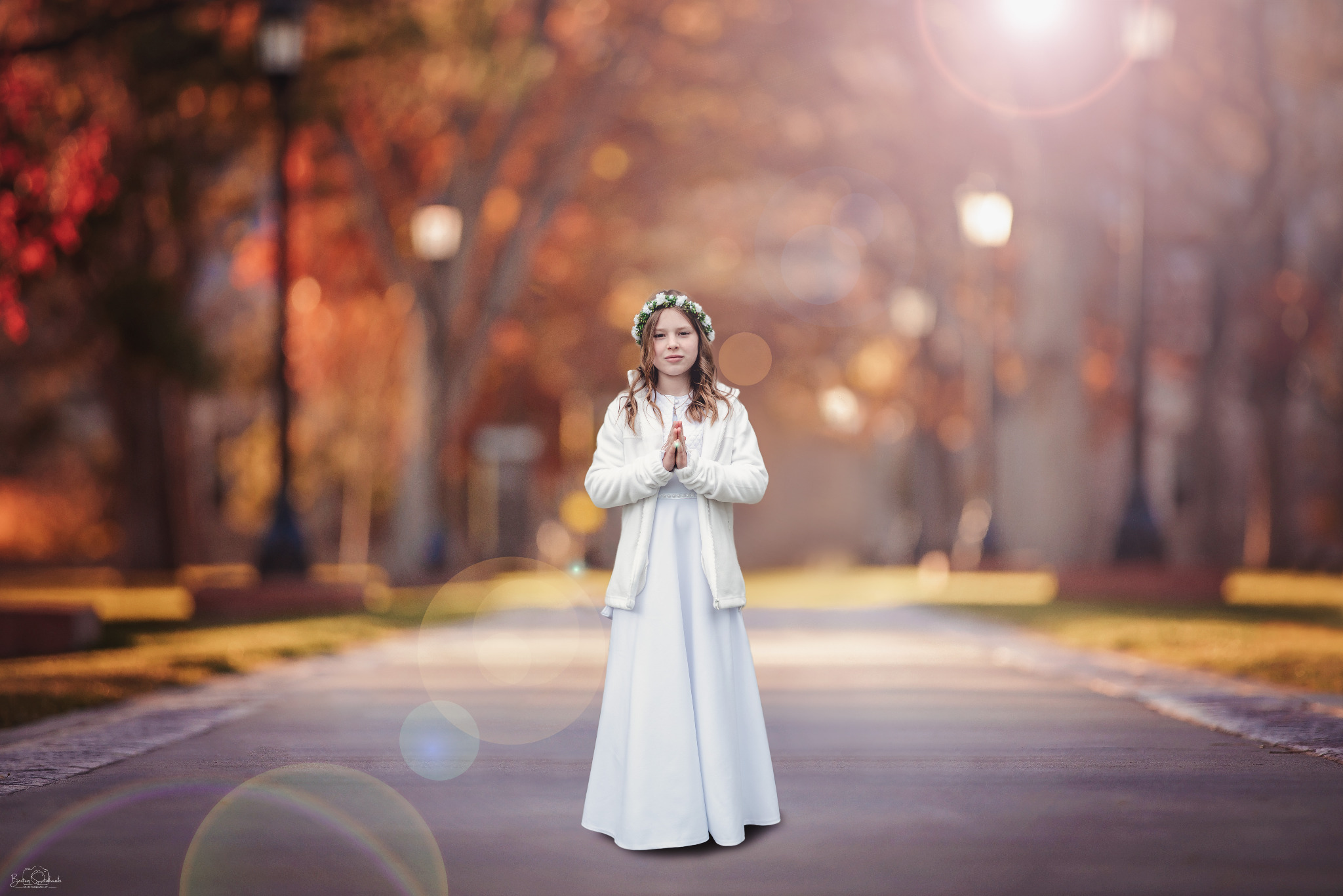 Dziewczynka w białej sukience i wianku na głowie, z rękami złożonymi do modlitwy, na tle jesiennego parku, w pełnym słońcu i z efektem bokeh.