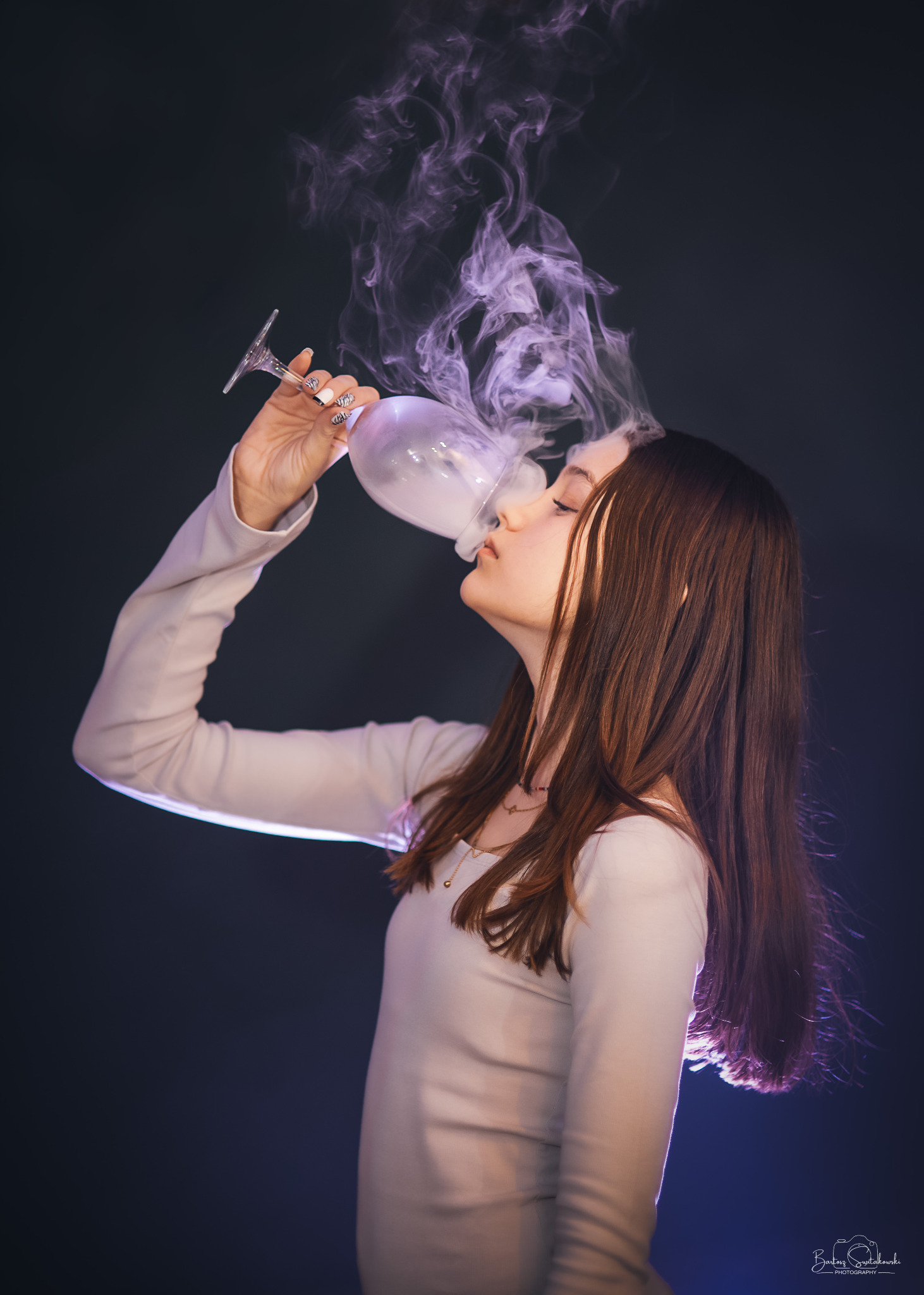 Profil kobiety pije dym z kieliszka na ciemnym tle. Wyrazisty manicure, długie włosy, delikatny makijaż. Efektowne oświetlenie i artystyczny klimat.