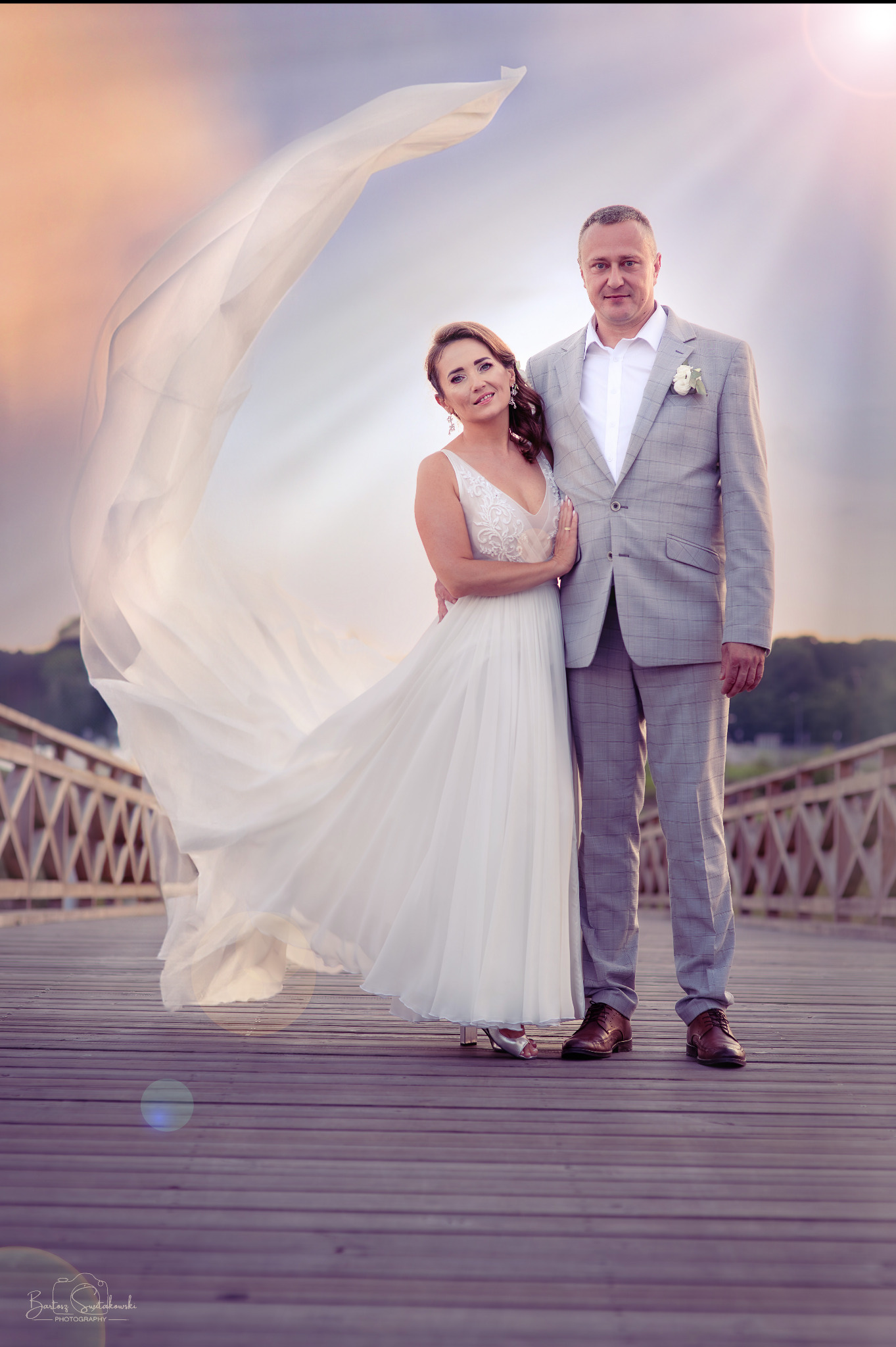 Para młoda na drewnianym pomoście w plenerze. Suknia z trenem powiewa na wietrze. Pan młody w szarej kracie. Romantyczna atmosfera, delikatne światło.